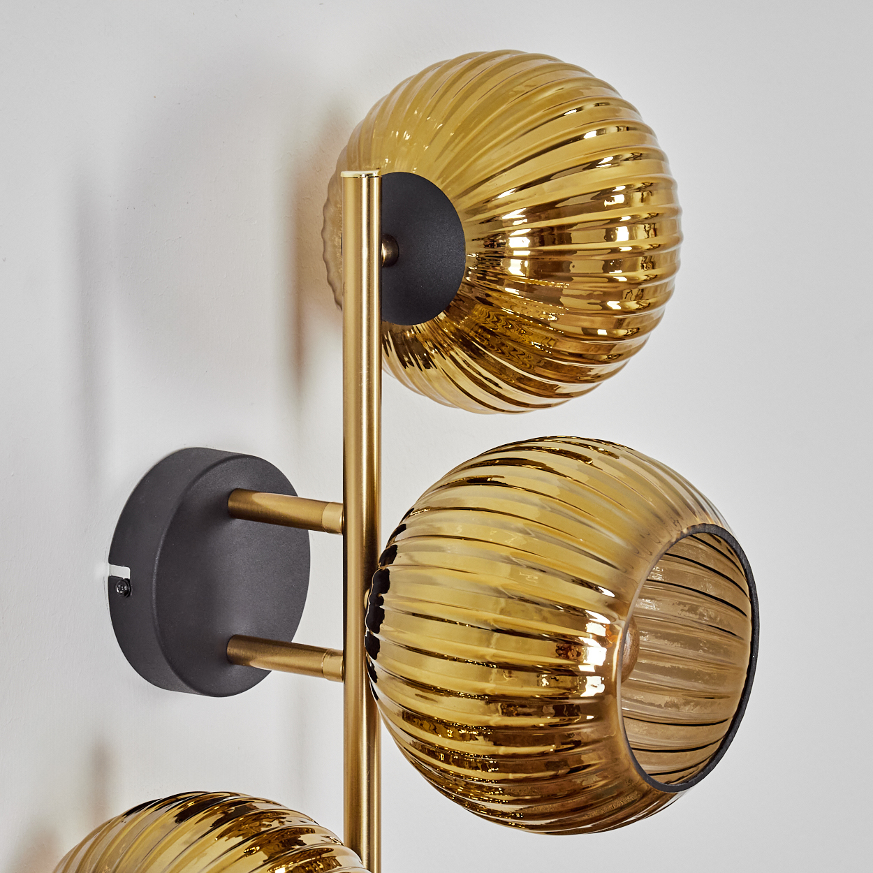 Eine Wandleuchte mit drei goldenen, gerippten Glaskugeln. Die Leuchte hat ein goldfarbenes Gestell und einen schwarzen Sockel an einer weißen Wand.