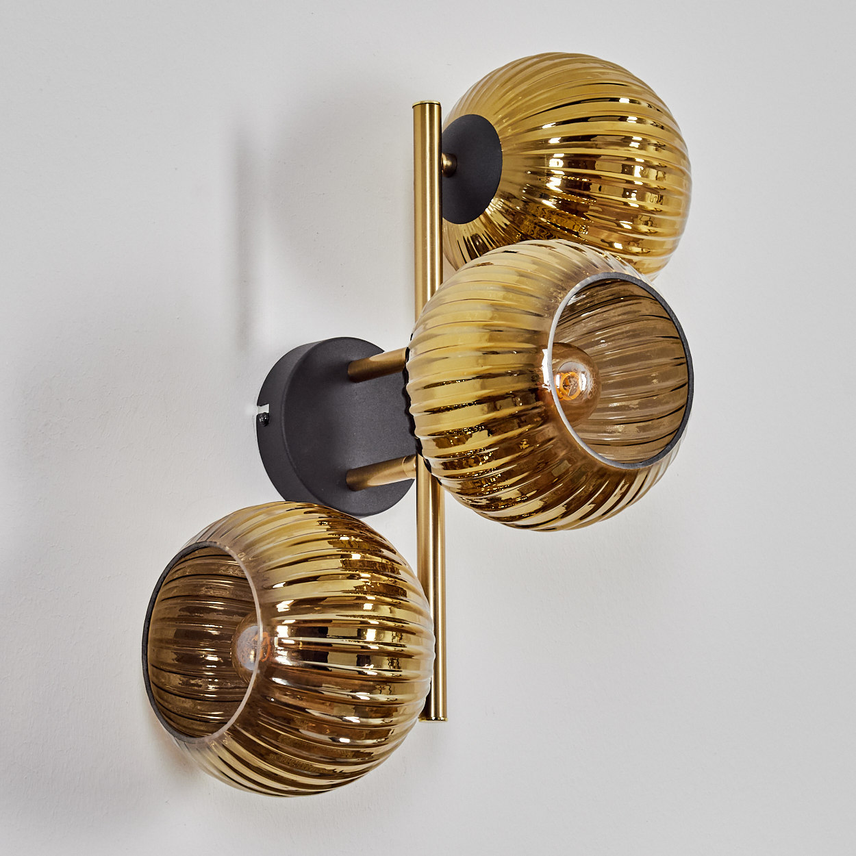 Eine Wandleuchte mit drei goldenen, gerippten Glaskugeln. Die Leuchte befindet sich vor einer weißen Wand.