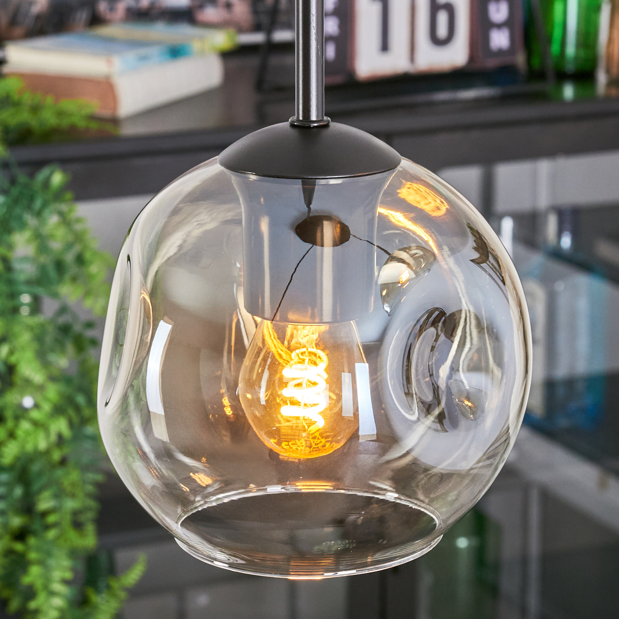 Eine moderne Glas-Pendelleuchte mit leuchtender Glühbirne im Inneren, hängend. Der Hintergrund umfasst grüne Pflanzen, Bücher und einen Kalender.