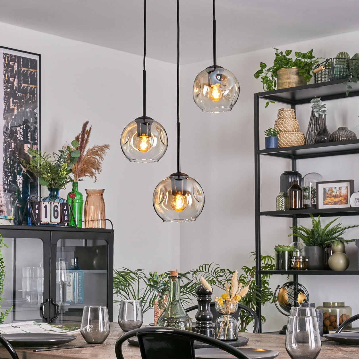 Esszimmer mit hängenden Kugelleuchten. Tisch mit Gläsern und Tellern gedeckt. Regal mit Pflanzen und Dekorationen im Hintergrund.