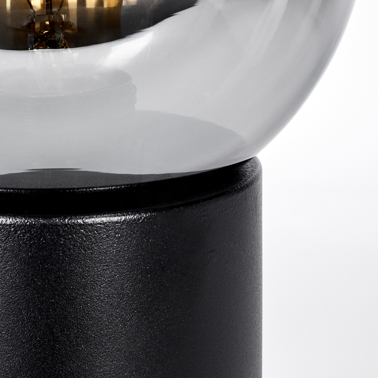 Nahaufnahme einer Lampe. Ein schwarzer, strukturierter Sockel trägt einen klaren, gebogenen Glasschirm. Der Glühfaden ist sichtbar.