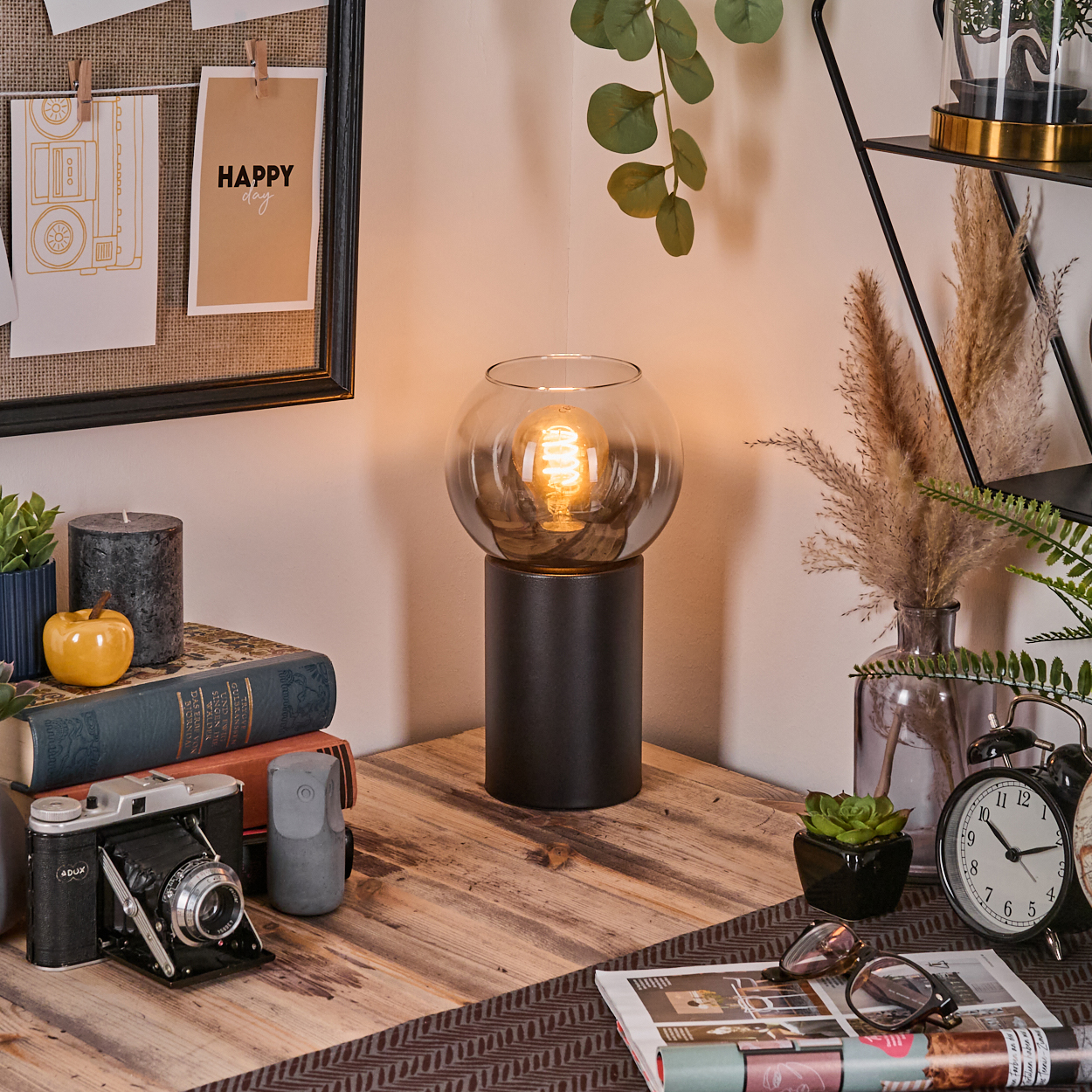 Ein Schreibtisch mit Lampe, Büchern, Kamera und Uhr. Eine Pinnwand mit Notizen hängt an der Wand. Pflanzen schmücken die Ecke.