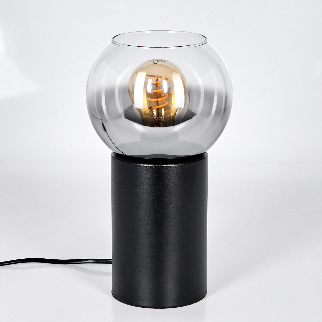 Eine moderne Lampe mit schwarzem zylindrischem Sockel und Glaskugelaufsatz. Eine Edison-Glühbirne befindet sich in der Glaskugel.