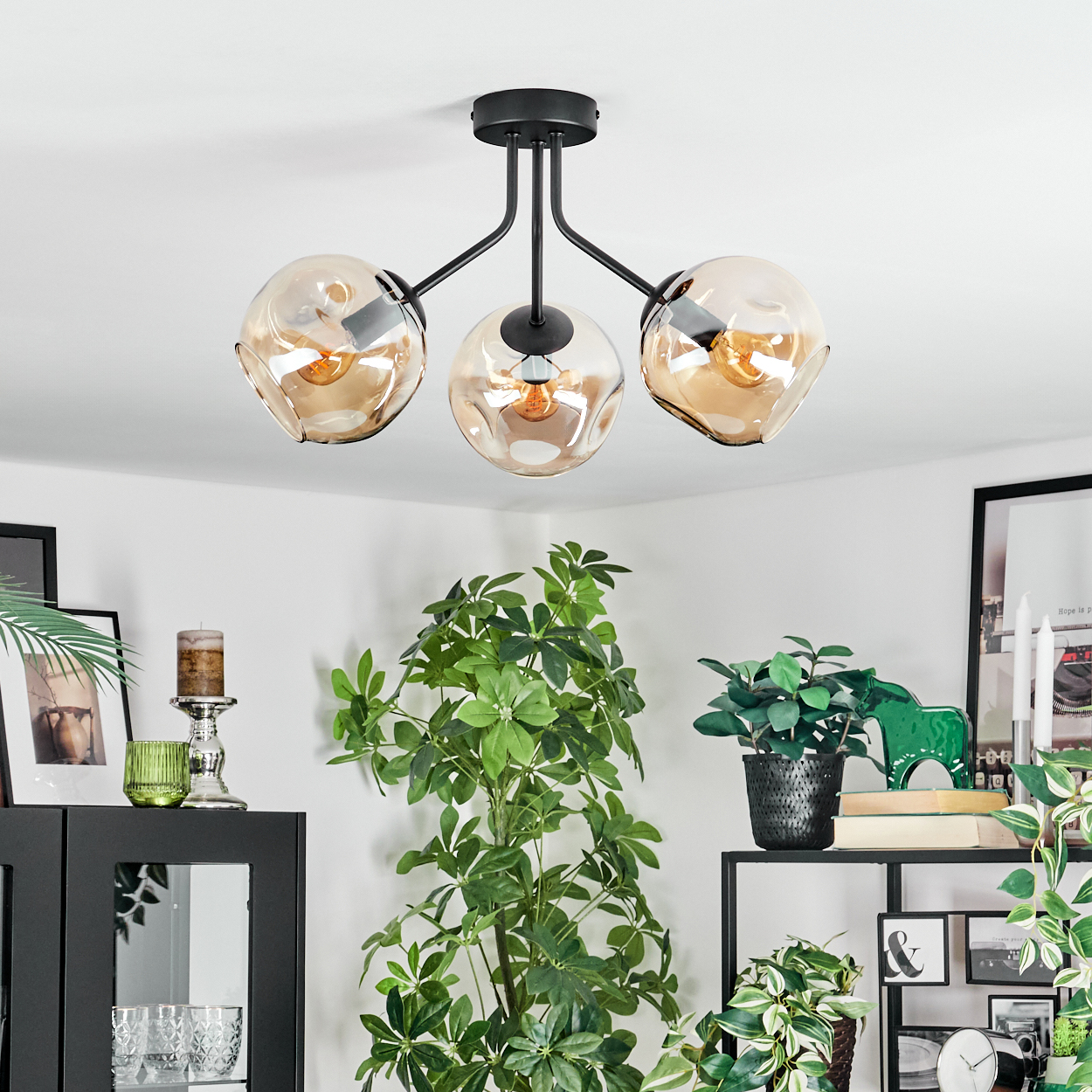 Eine moderne Deckenlampe mit drei bernsteinfarbenen Glasschirmen. Darunter grüne Pflanzen, schwarze Regale und Dekorationsartikel.