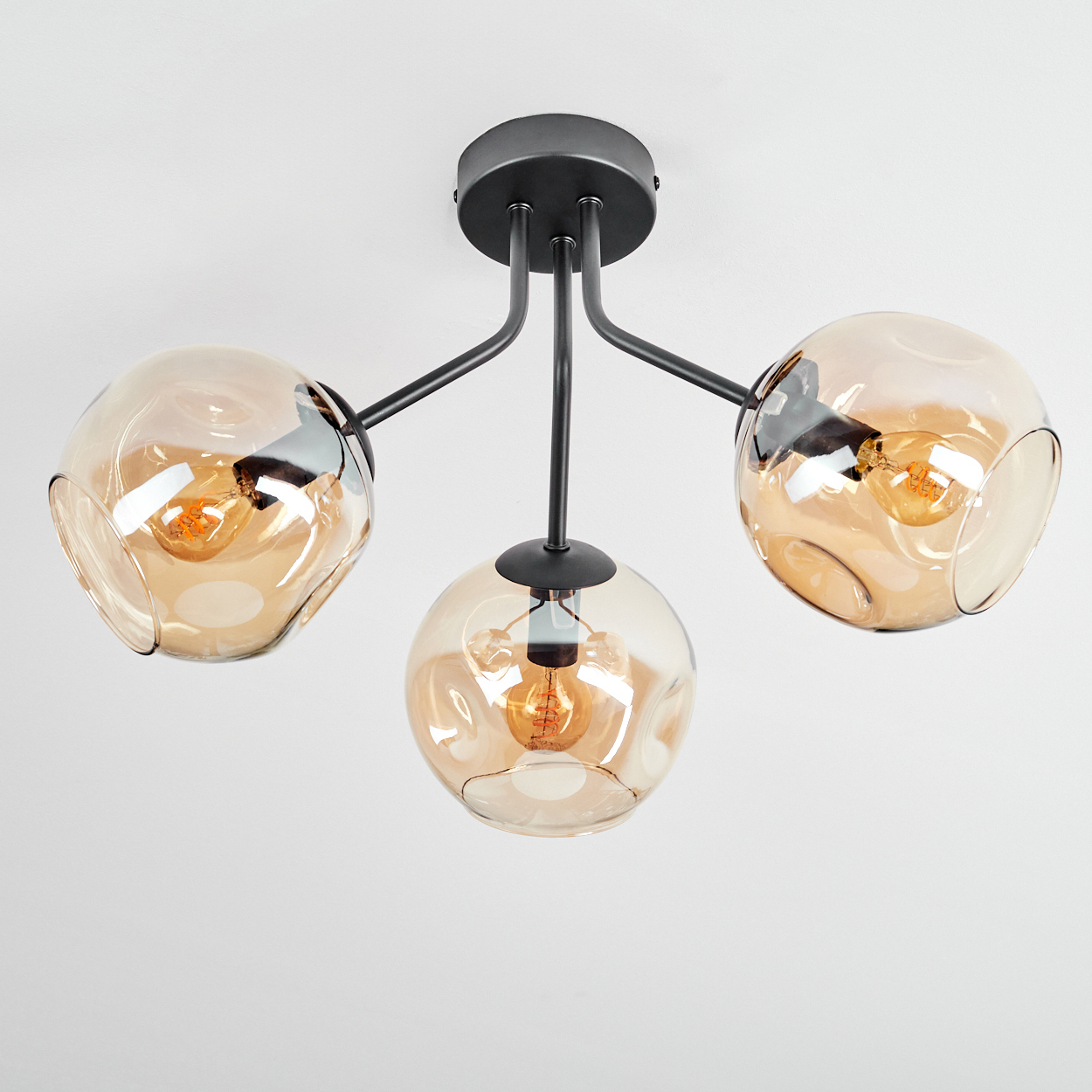Moderne Deckenleuchte mit drei bernsteinfarbenen Glasschirmen und schwarzen Metallarmen, jeweils mit Glühbirne. Der Hintergrund ist weiß.