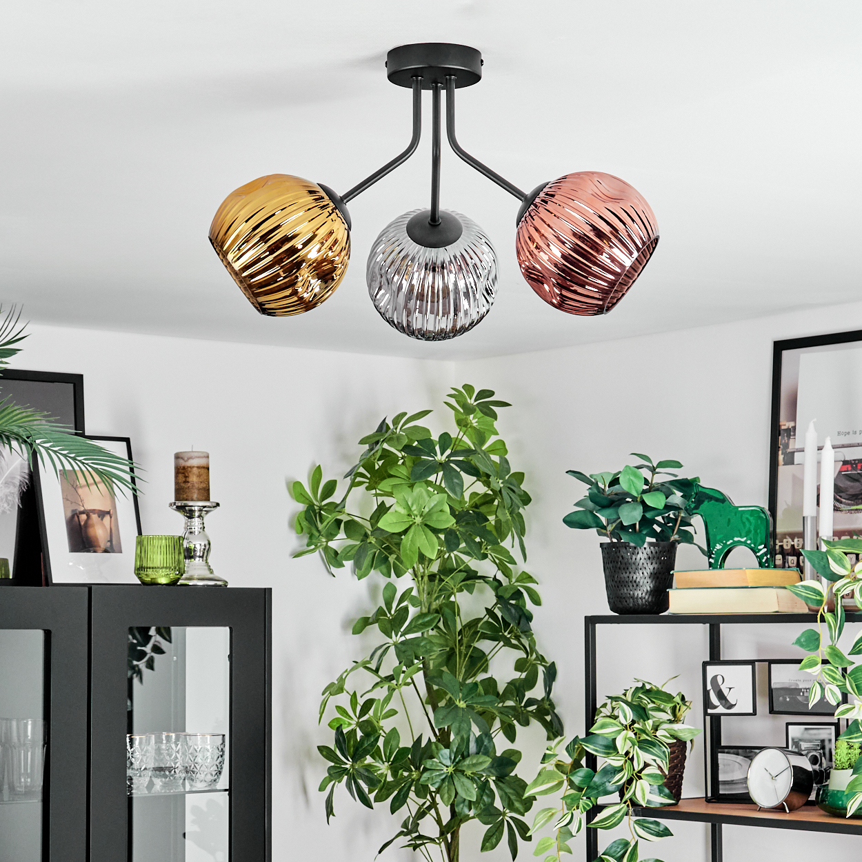 Eine moderne Deckenleuchte mit drei Glaskugeln in Gold, Silber und Kupfer. Dekorative Pflanzen und Regale mit verschiedenen Gegenständen im Hintergrund.