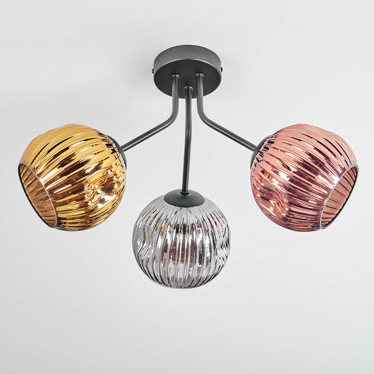 Deckenlampe mit drei kugelförmigen Schirmen. Einer gold, einer silber und einer kupferfarben. Die Halterung ist schwarz, an weißer Decke montiert.