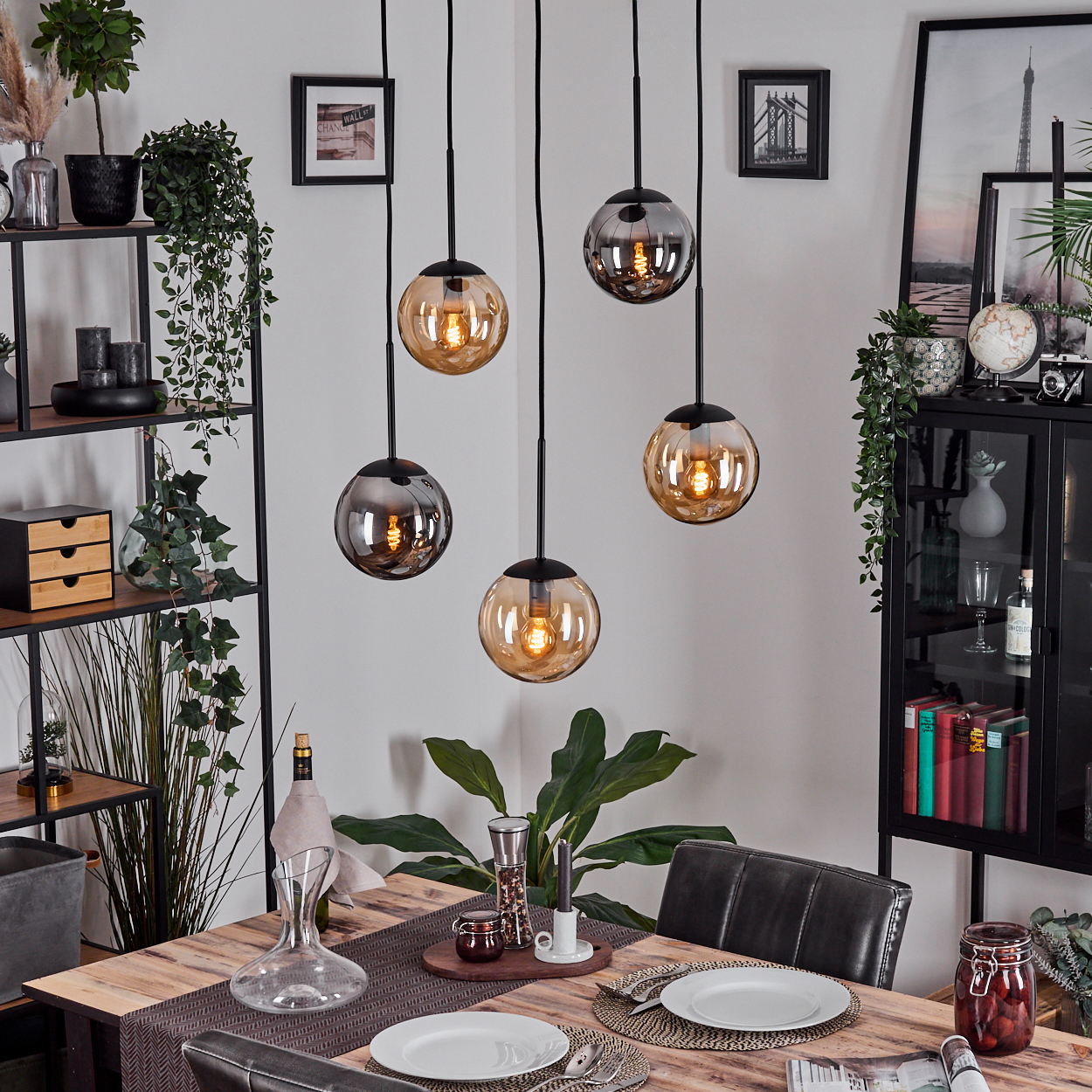 Esszimmer mit Holztisch. Hängelampen hängen darüber. Ein Schrank, Regale, Pflanzen und eine Karaffe sind sichtbar.