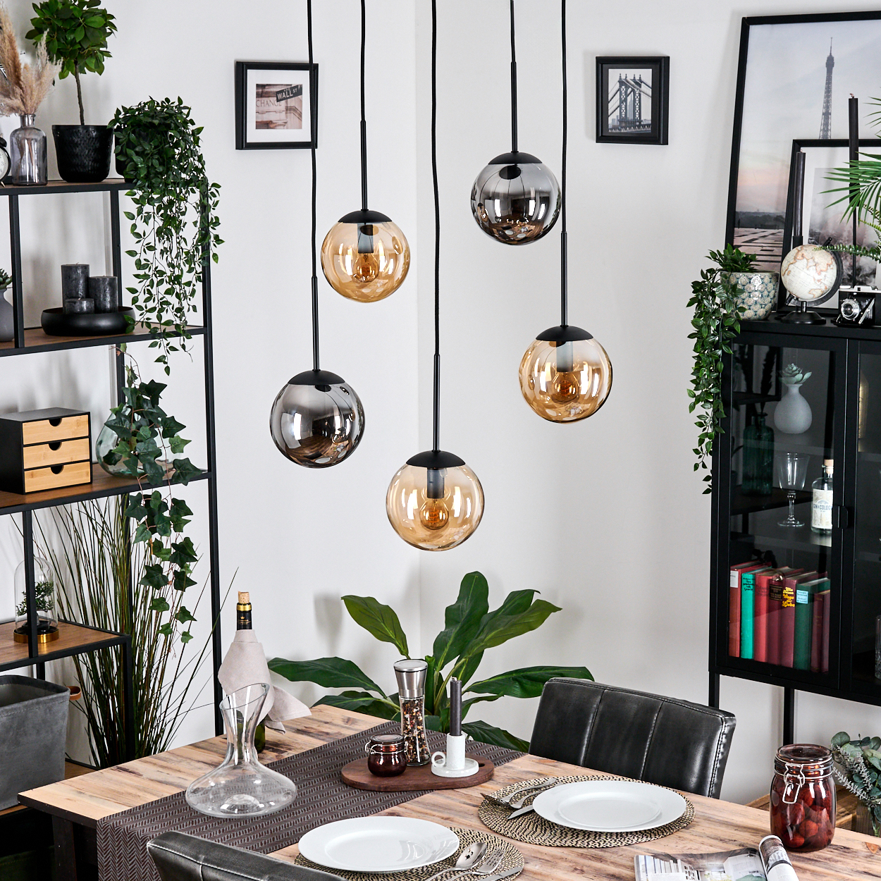 Esszimmer mit Holztisch, Hängelampen, Pflanzen und Dekoration. Enthält Teller, Gläser und eine Glaskaraffe.
