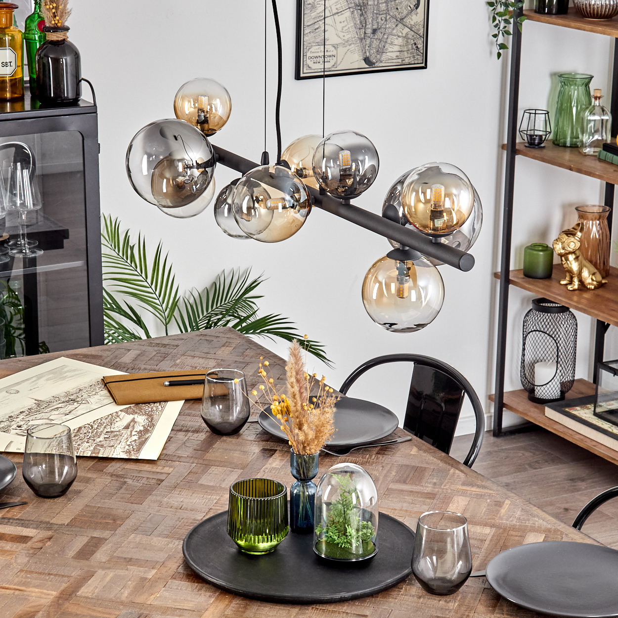 Ein Esstisch mit Gläsern, Tellern und Dekorationen. Oben eine moderne Deckenleuchte mit kugelförmigen Lichtern. Regale mit Gegenständen sind im Hintergrund.