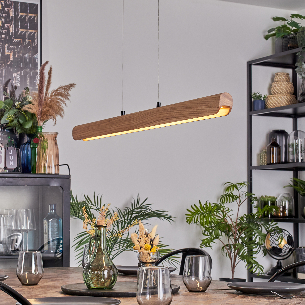 Hölzerne Lampe hängt über einem Esstisch. Tisch gedeckt mit Gläsern und Tellern. Pflanzen und Dekorationen im Hintergrund.