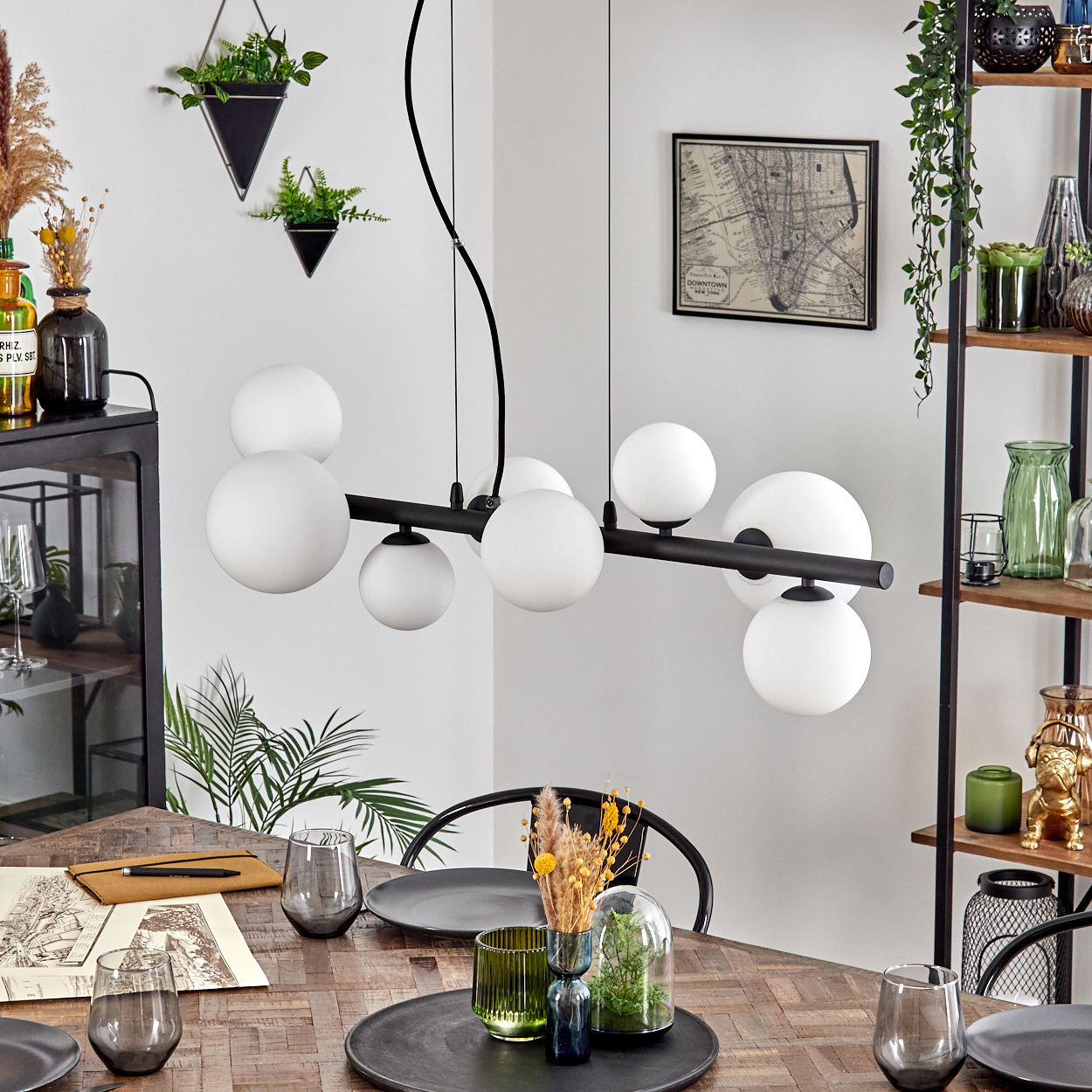 Esszimmer mit Holztisch, schwarzen Stühlen und modernem Kronleuchter. Dekoration umfasst Pflanzen, Gläser und eine Karte.