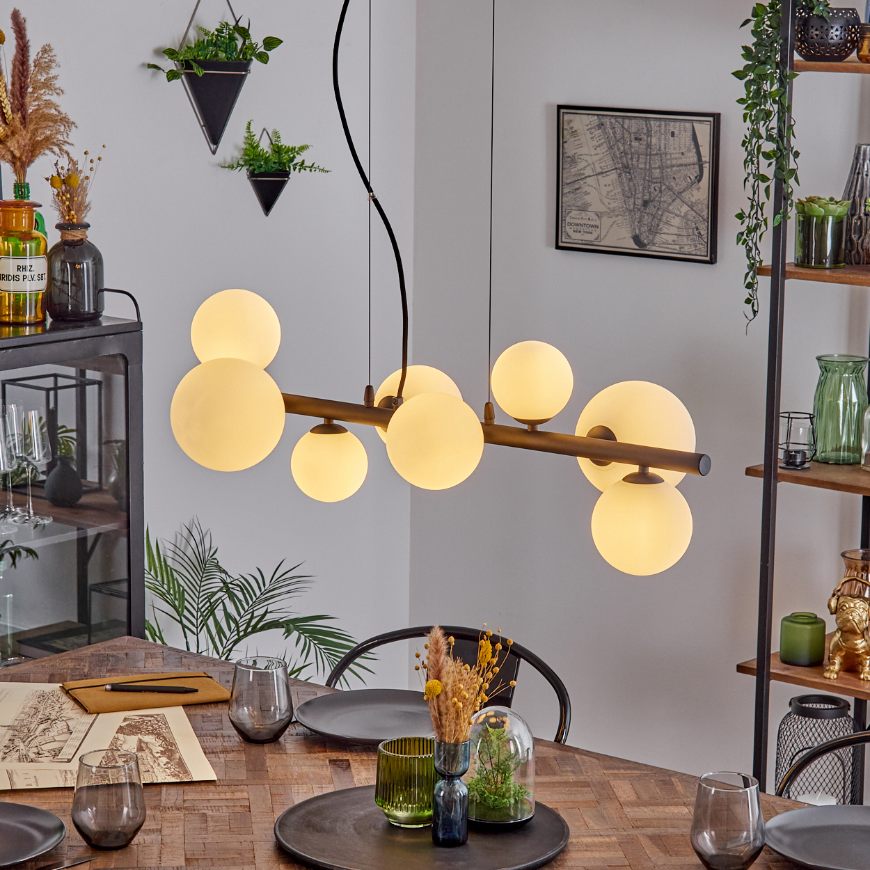 Esszimmer mit Holztisch, modernem Kronleuchter mit Kugellichtern und dekorativen Pflanzen. Eine Karte hängt an der Wand.