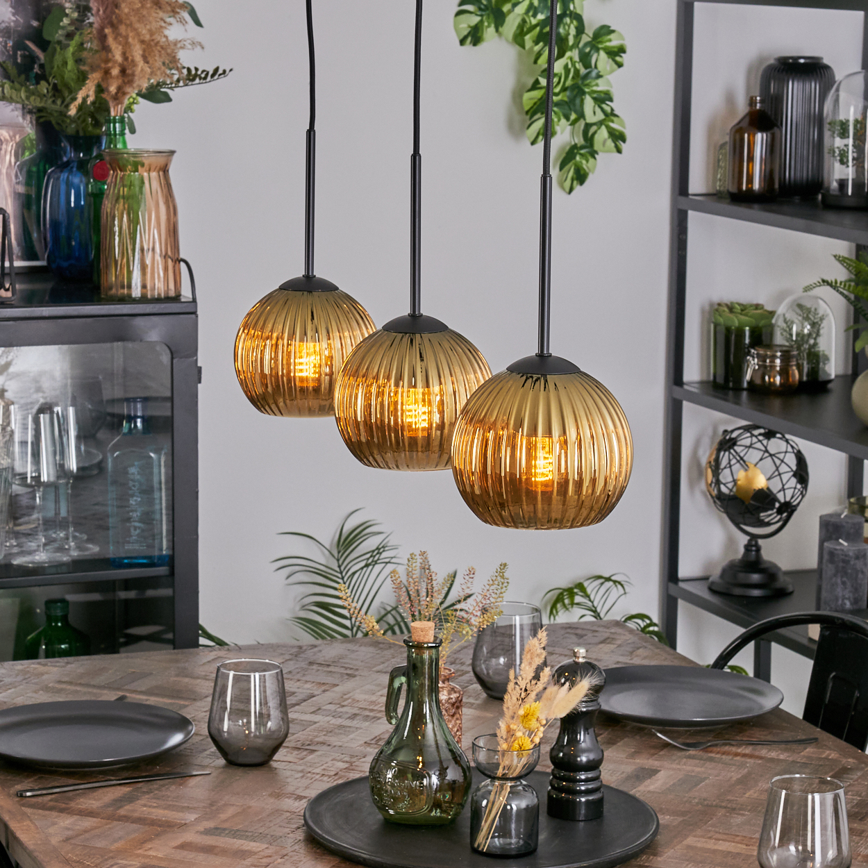 Drei goldene Pendelleuchten hängen über einem Holztisch, der mit Tellern, Gläsern und Dekoration gedeckt ist. Im Hintergrund ein Regal mit Dekoration.