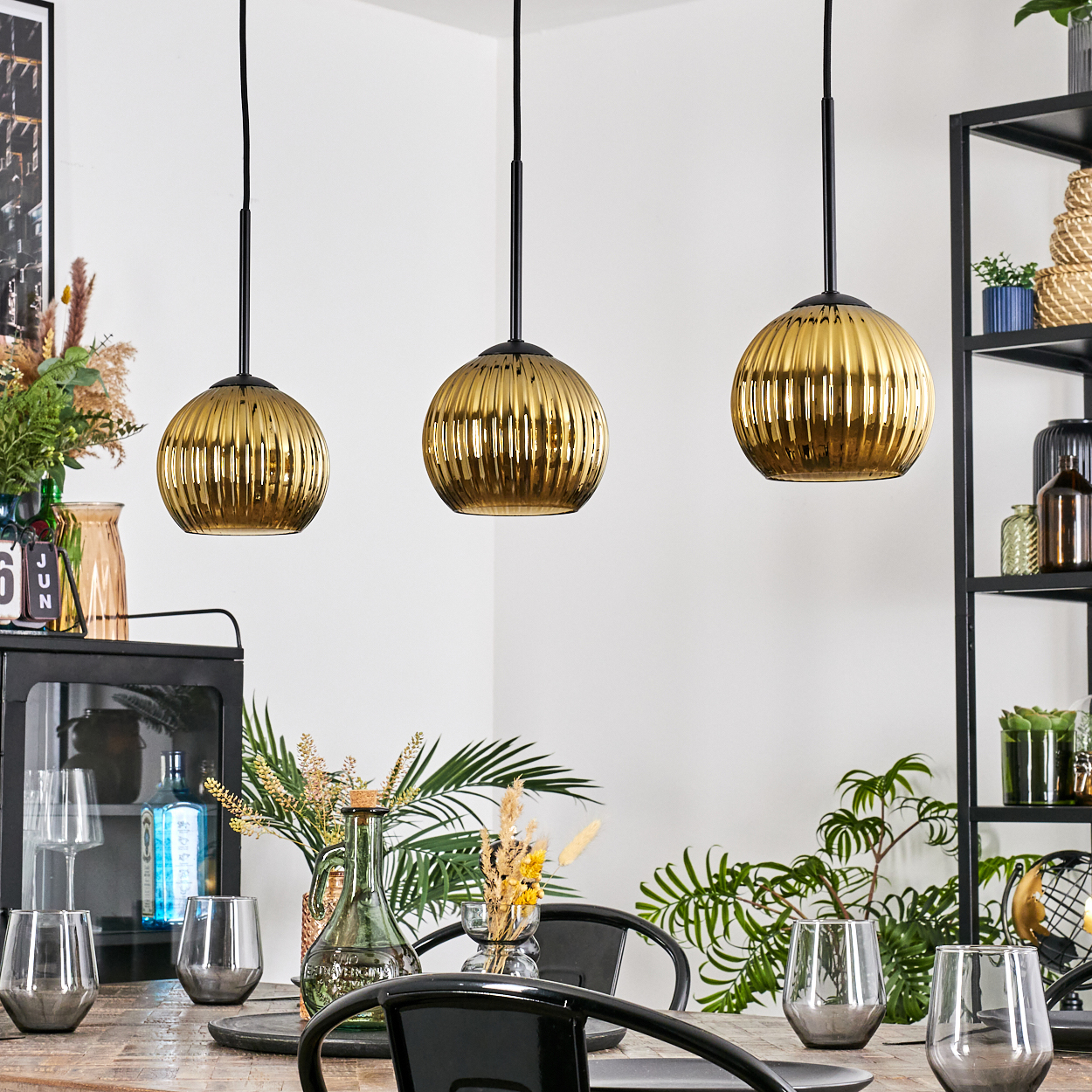 Drei goldene Pendelleuchten hängen über einem Holztisch, der mit Gläsern und Dekoration gedeckt ist. Pflanzen und ein schwarzes Regal sind sichtbar.