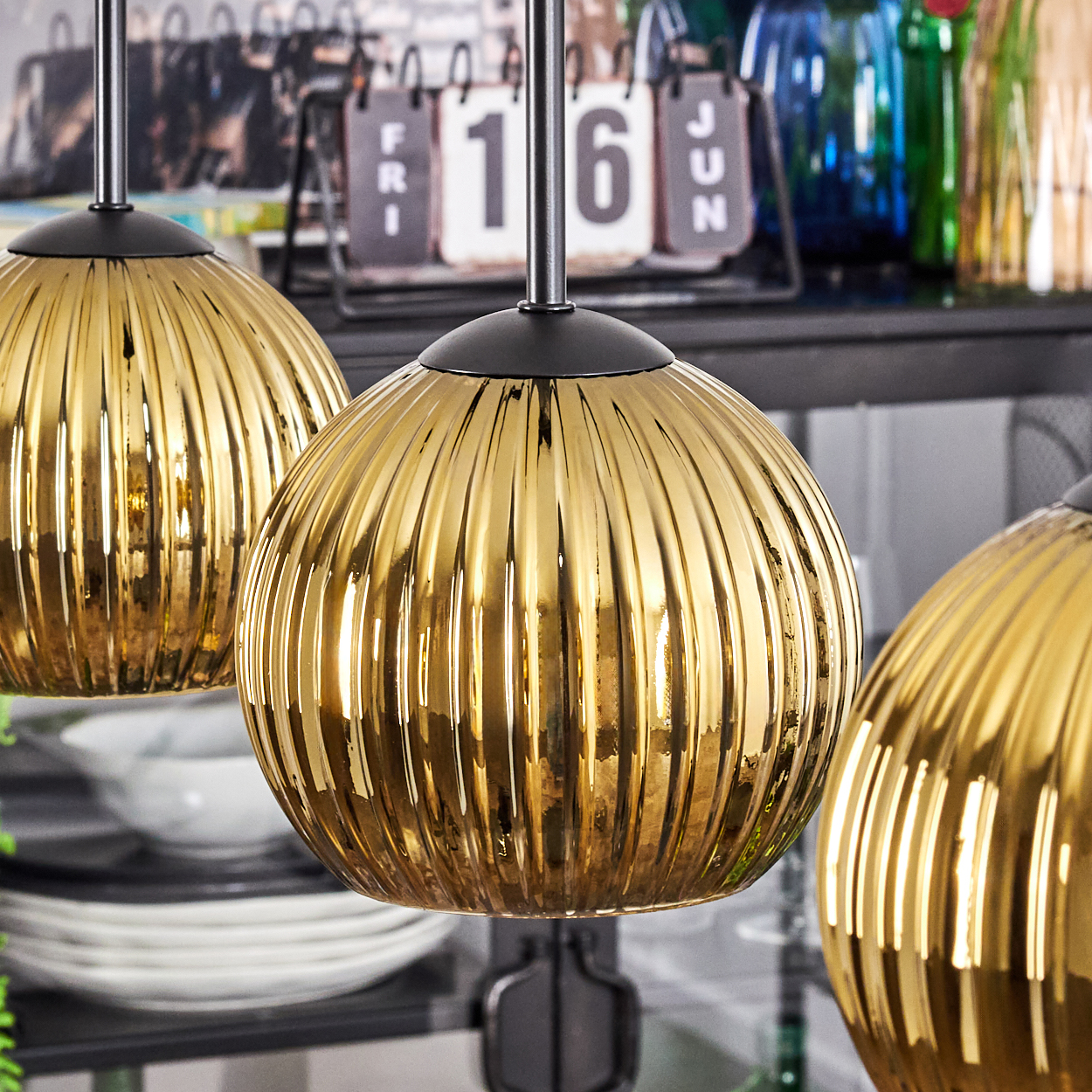 Drei goldene, gerippte Glas-Hängelampen hängen. Ein Kalender zeigt die 16. Geschirr und Flaschen sind im Hintergrund.