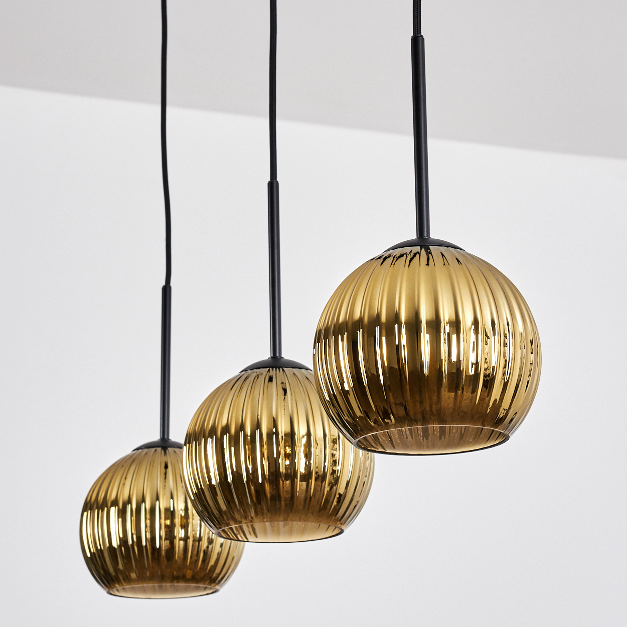 Drei goldene, geriffelte Pendelleuchten hängen von der Decke. Schwarze Kabel und Stiele stützen die kugelförmigen Glasschirme.