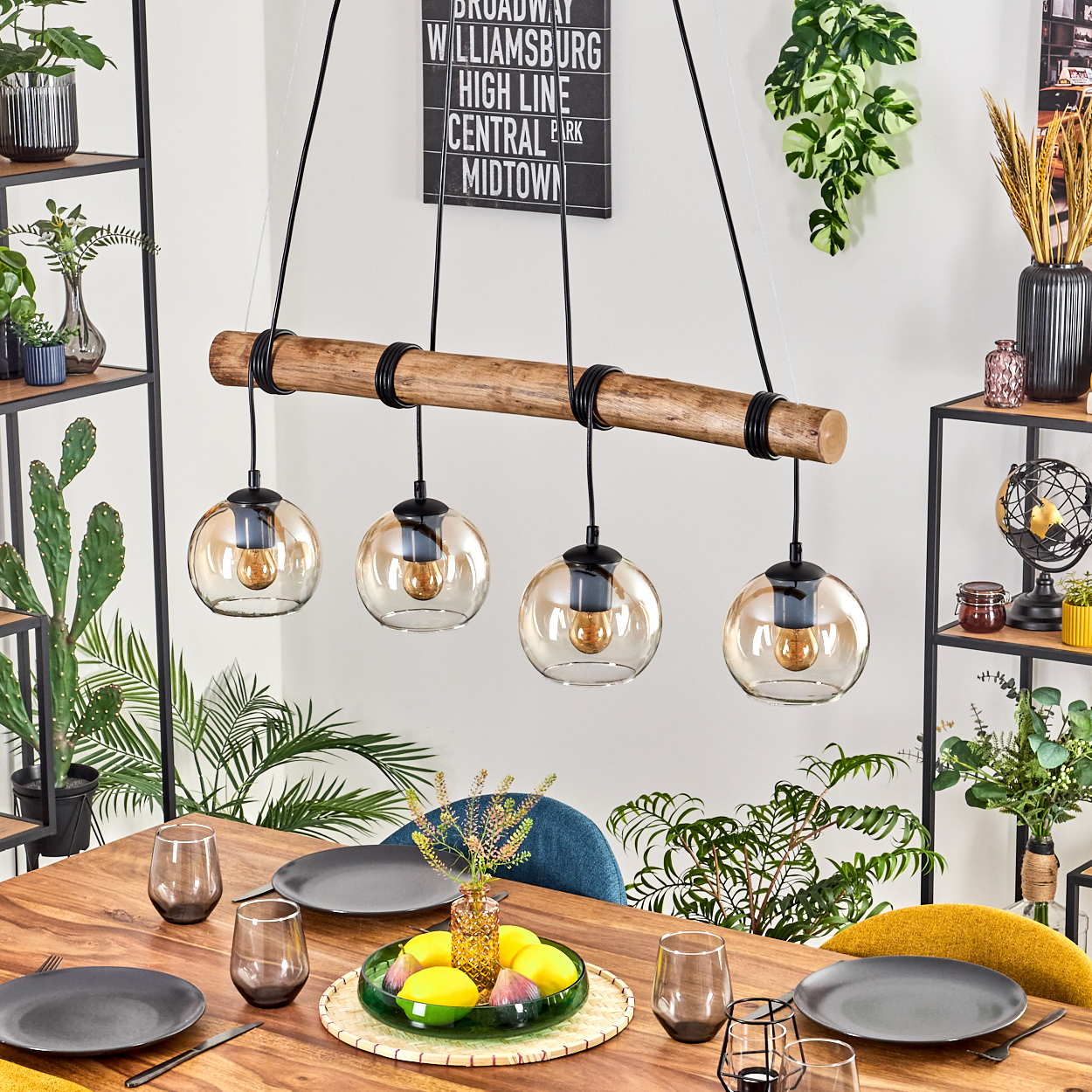 Ein Esszimmer mit Holztisch, Hängelampen und Pflanzen. Teller, Gläser und eine Obstschale stehen auf dem Tisch.