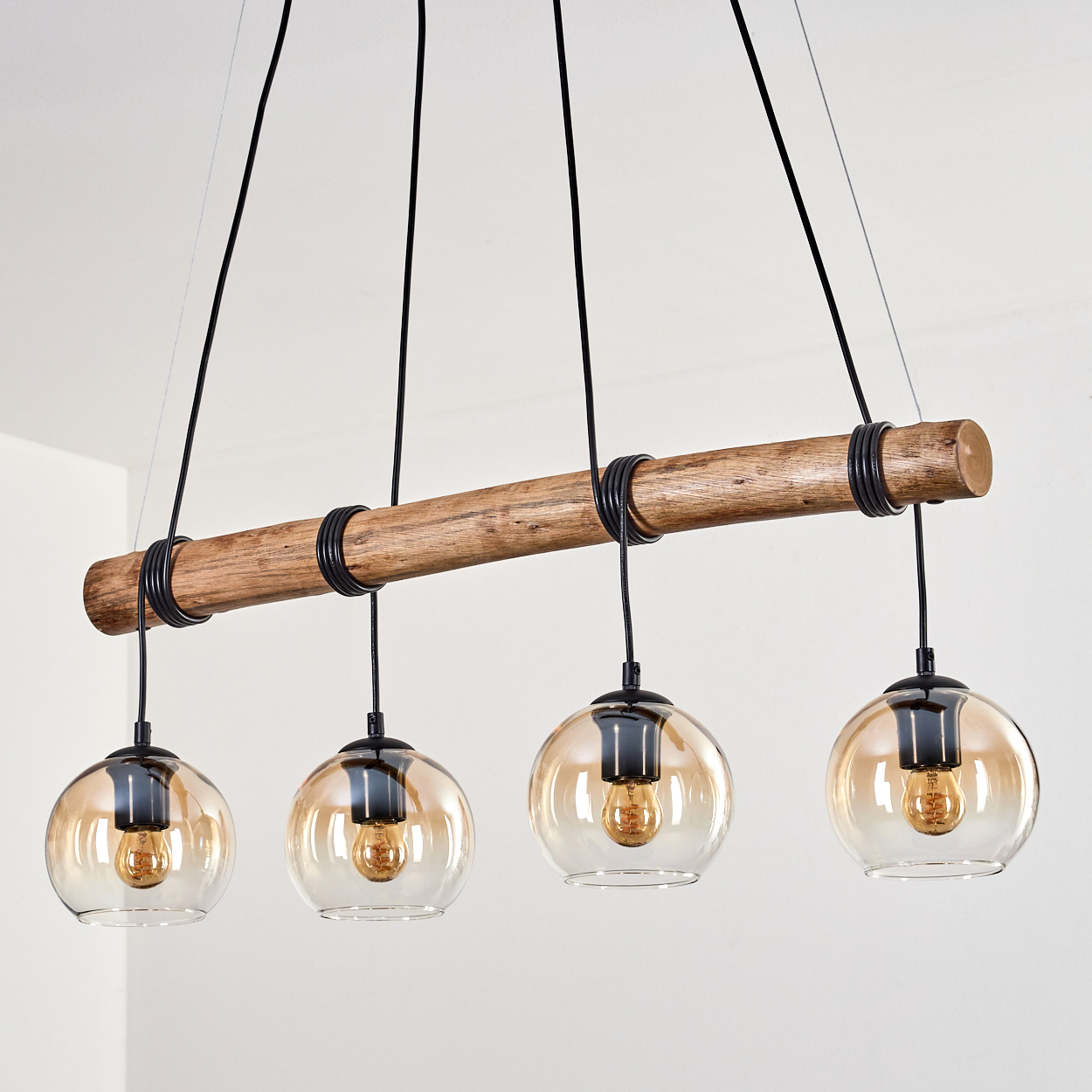 Eine Holzbalken-Kronleuchter mit vier Glaskugellampen, die an schwarzen Drähten aufgehängt sind. Der Hintergrund ist eine weiße Wand.