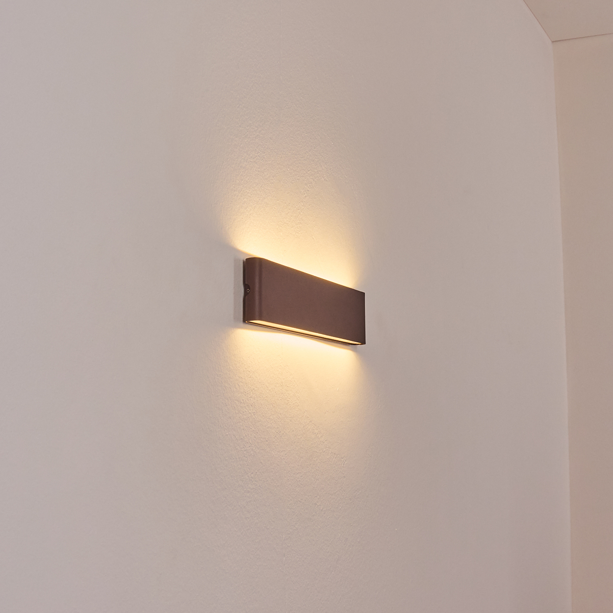 Eine rechteckige, bronzefarbene Wandleuchte strahlt warmes Licht aus und wirft Schatten auf eine weiße Wand. Die Lampe ist an der Wand montiert.