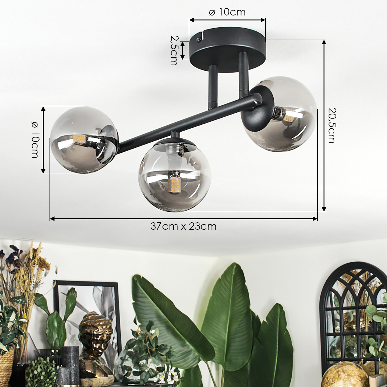 Schwarze Deckenlampe mit drei kugelförmigen Schirmen. Abmessungen sind angegeben. Vordergrund hat Pflanzen und Dekor.