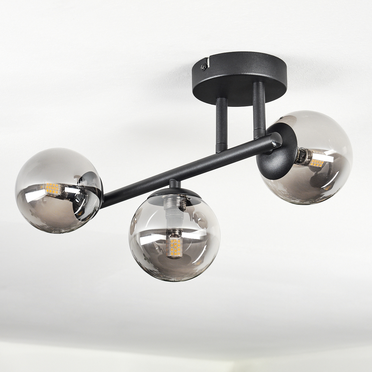 Moderne Deckenleuchte mit drei rauchfarbenen Glaskugeln und einem schwarzen Metallrahmen, vor weißer Decke.