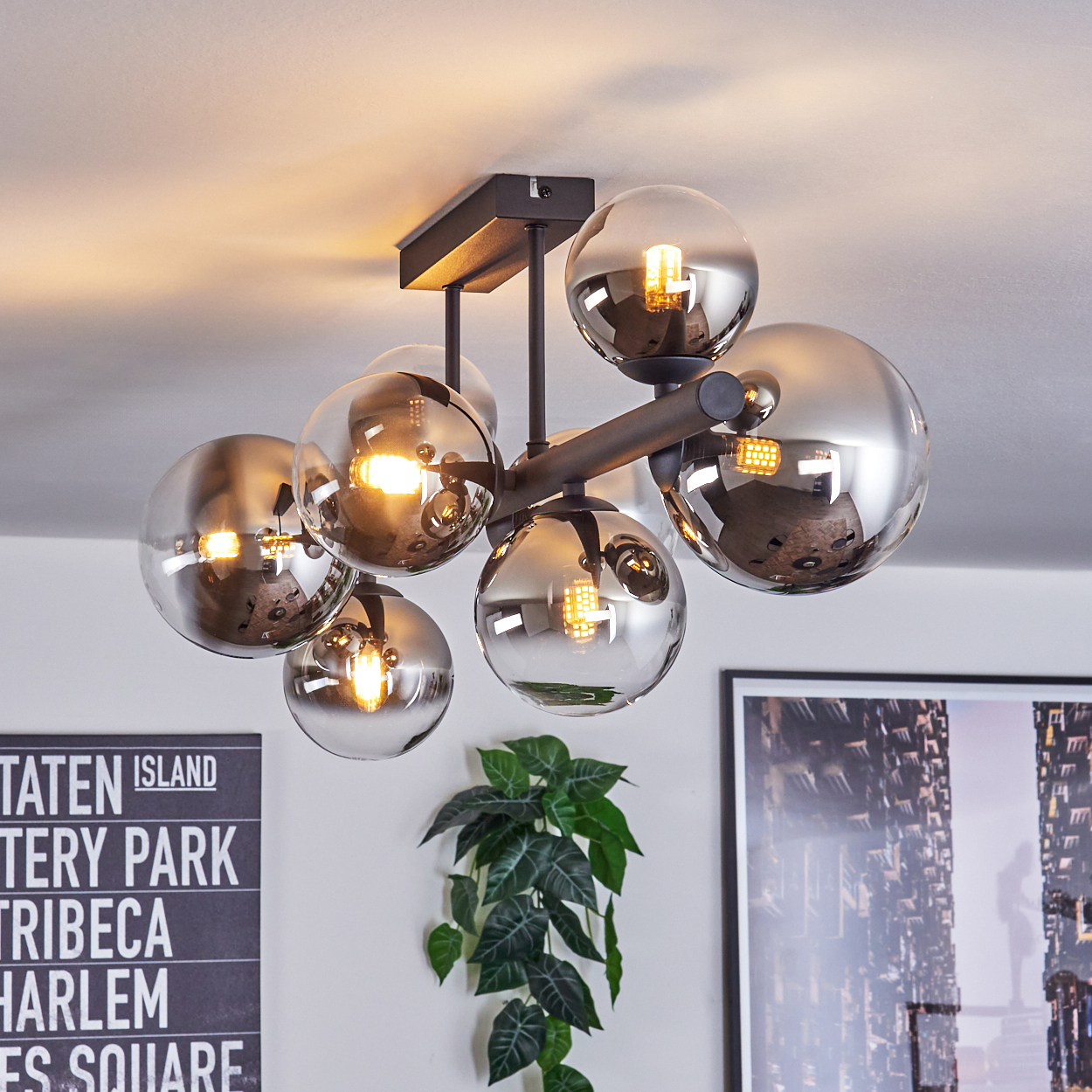 Eine moderne Deckenleuchte mit mehreren kugelförmigen Glasschirmen. Die Leuchte hängt von der Decke. Eine Pflanze und Kunstwerke sind sichtbar.