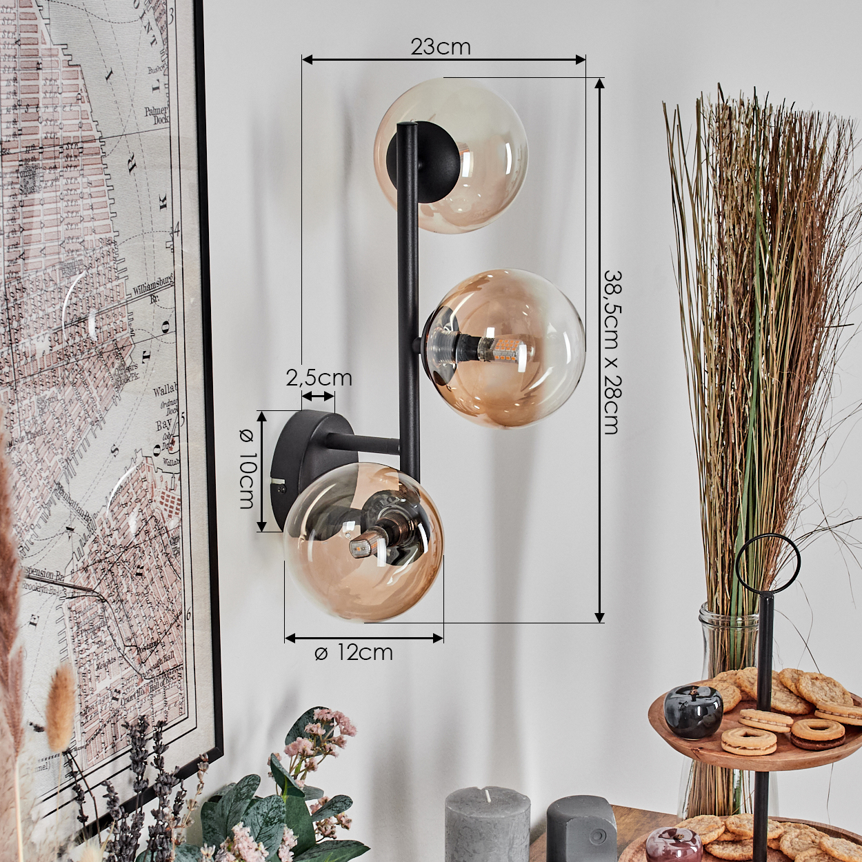 Eine moderne Wandlampe mit drei Glaskugeln. Sie ist an einer weißen Wand montiert. Weitere Dekorationen sind eine Karte, Trockengras und Kekse.