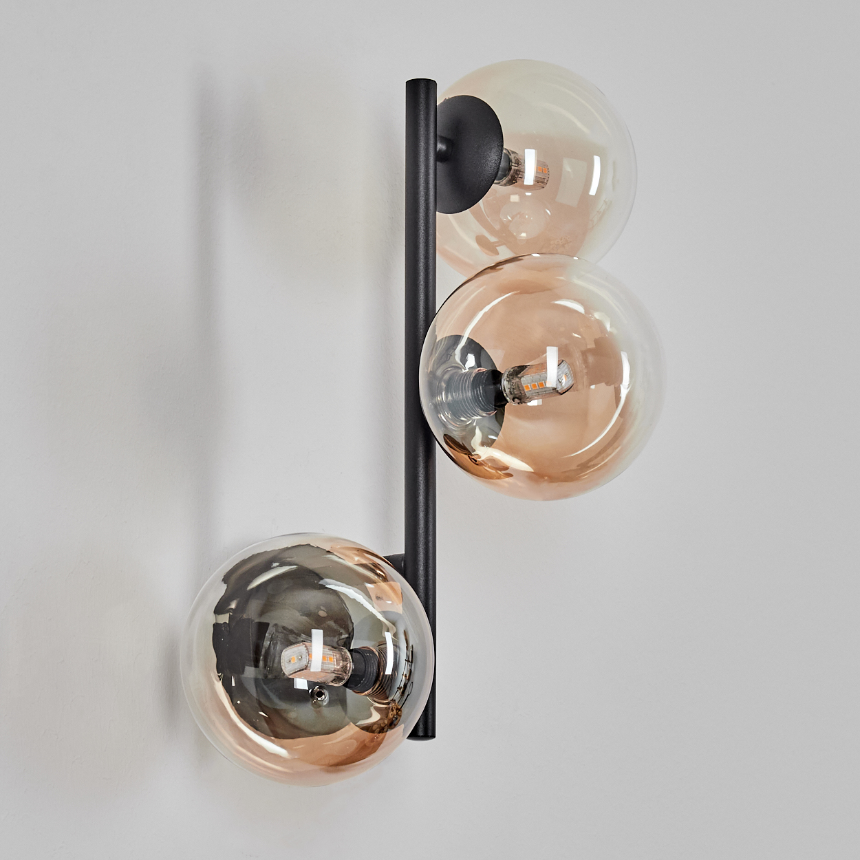 Moderne Wandlampe mit drei kugelförmigen Leuchten. Die Schirme sind bernsteinfarben, der Sockel schwarz an einer weißen Wand.