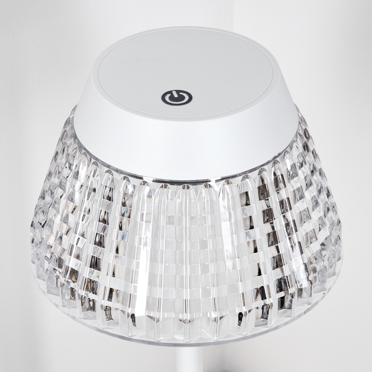 Eine weiße und klare Lampe steht auf einer weißen Oberfläche. Die Lampe hat oben einen Ein-/Ausschalter. Der Lampenschirm ist klar und strukturiert.
