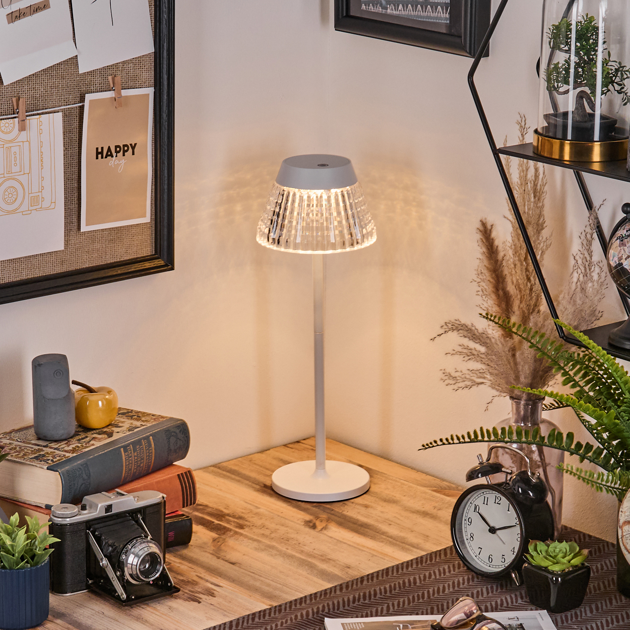 Eine beleuchtete Lampe auf einem Holztisch mit Büchern, einer Kamera und einer Uhr. Eine Pinnwand mit einem 'Happy Day'-Zettel im Hintergrund.