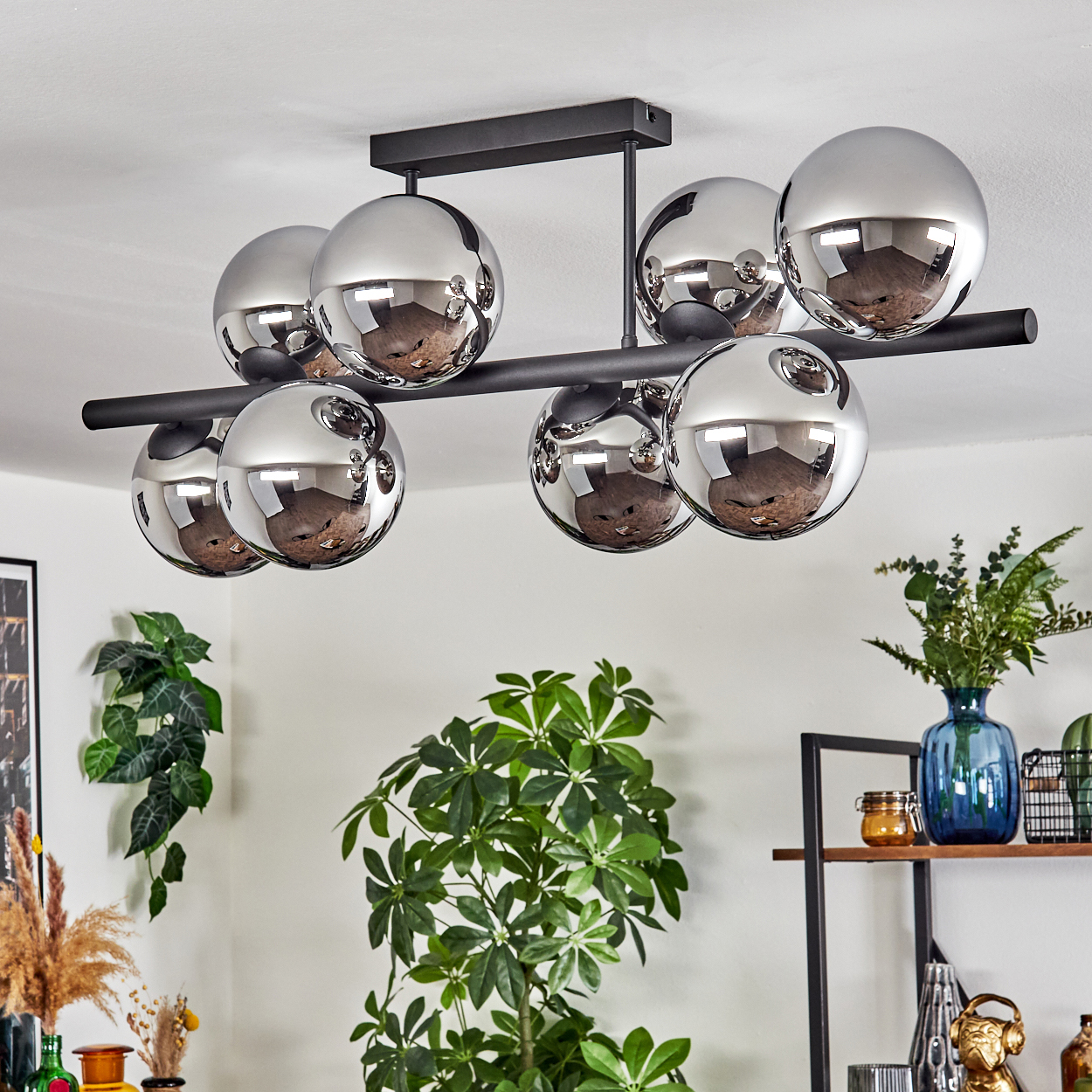 Eine moderne Deckenleuchte mit Chromkugeln. Darunter sind Zimmerpflanzen und ein Regal mit Dekoration zu sehen, was eine gemütliche Atmosphäre schafft.