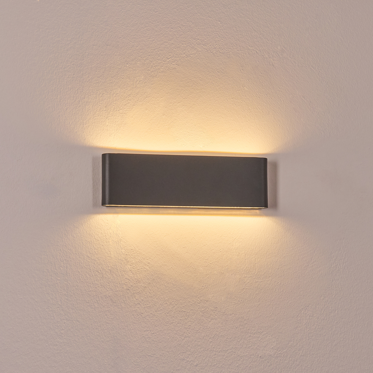 Eine dunkelgraue, rechteckige Wandleuchte ist an einer strukturierten weißen Wand befestigt. Die Lampe strahlt ein warmes, weiches Licht aus.