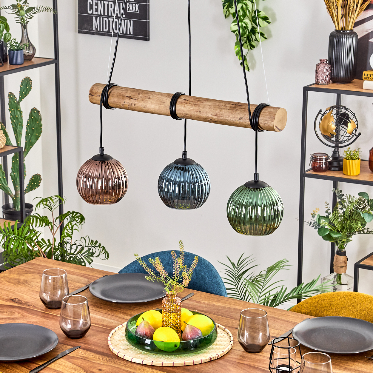Esszimmer mit Holztisch, drei Hängelampen, Tellern, Gläsern und einer Obstschale. Pflanzen und Regale sind sichtbar.