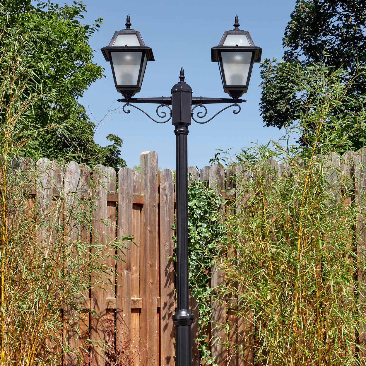 Eine schwarze Laterne mit zwei Lampen steht vor einem Holzzaun. Bambus und grünes Laub säumen die Säule vor blauem Himmel.