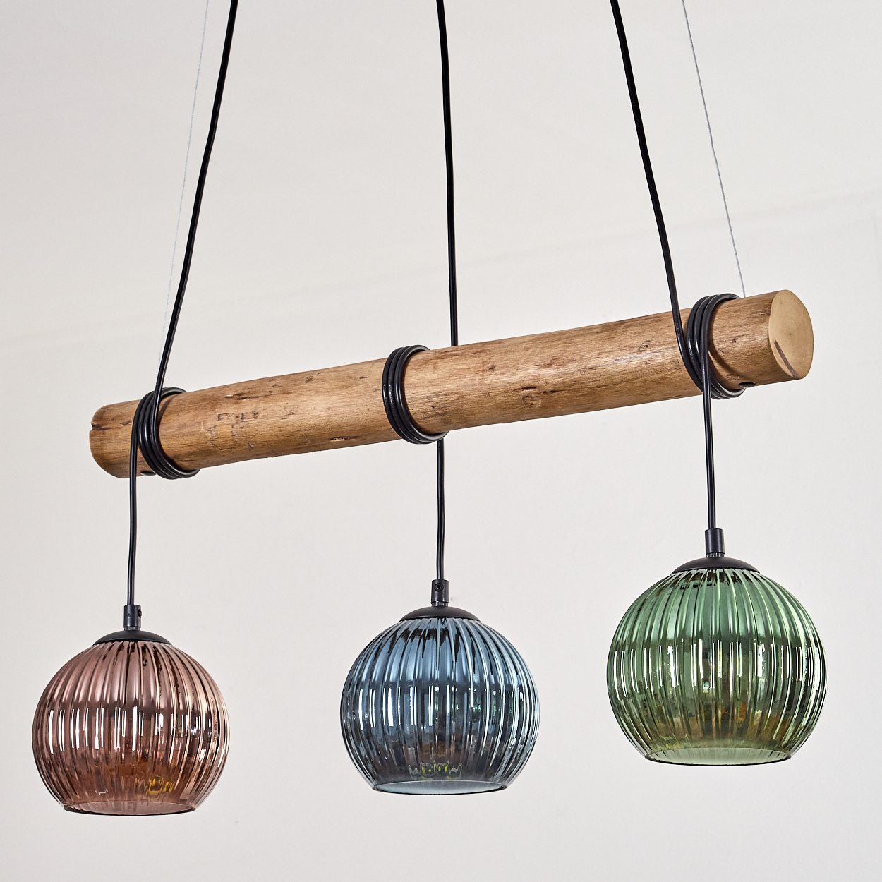 Drei Pendelleuchten mit Glaskugeln in Rosa, Blau und Grün hängen an einem Holzbalken. Schwarze Drähte halten die Lampen.