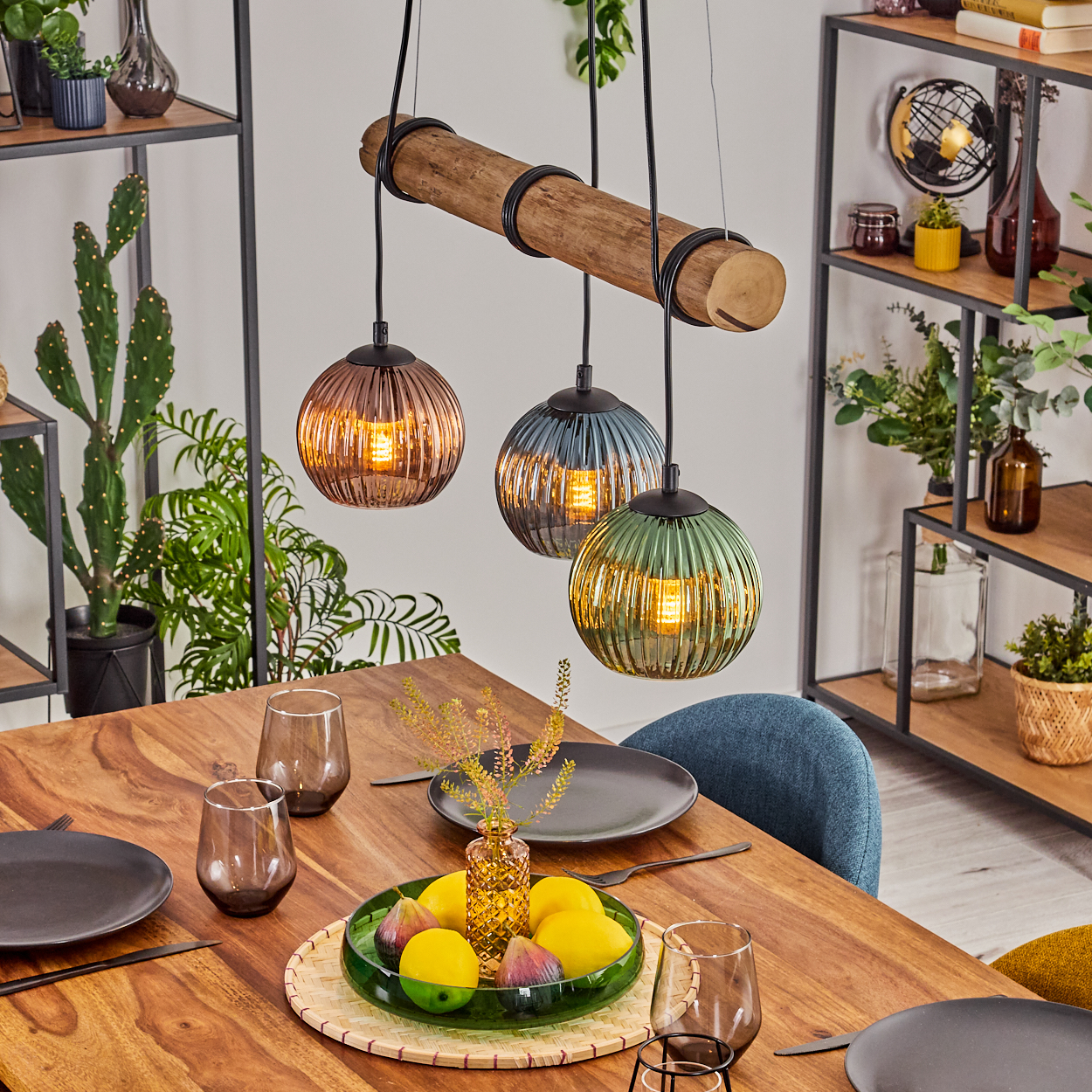 Ein Esstisch mit Holzplatte, gedeckt mit Tellern, Gläsern, Obst und beleuchteten Pendelleuchten. Regale mit Deko im Hintergrund.