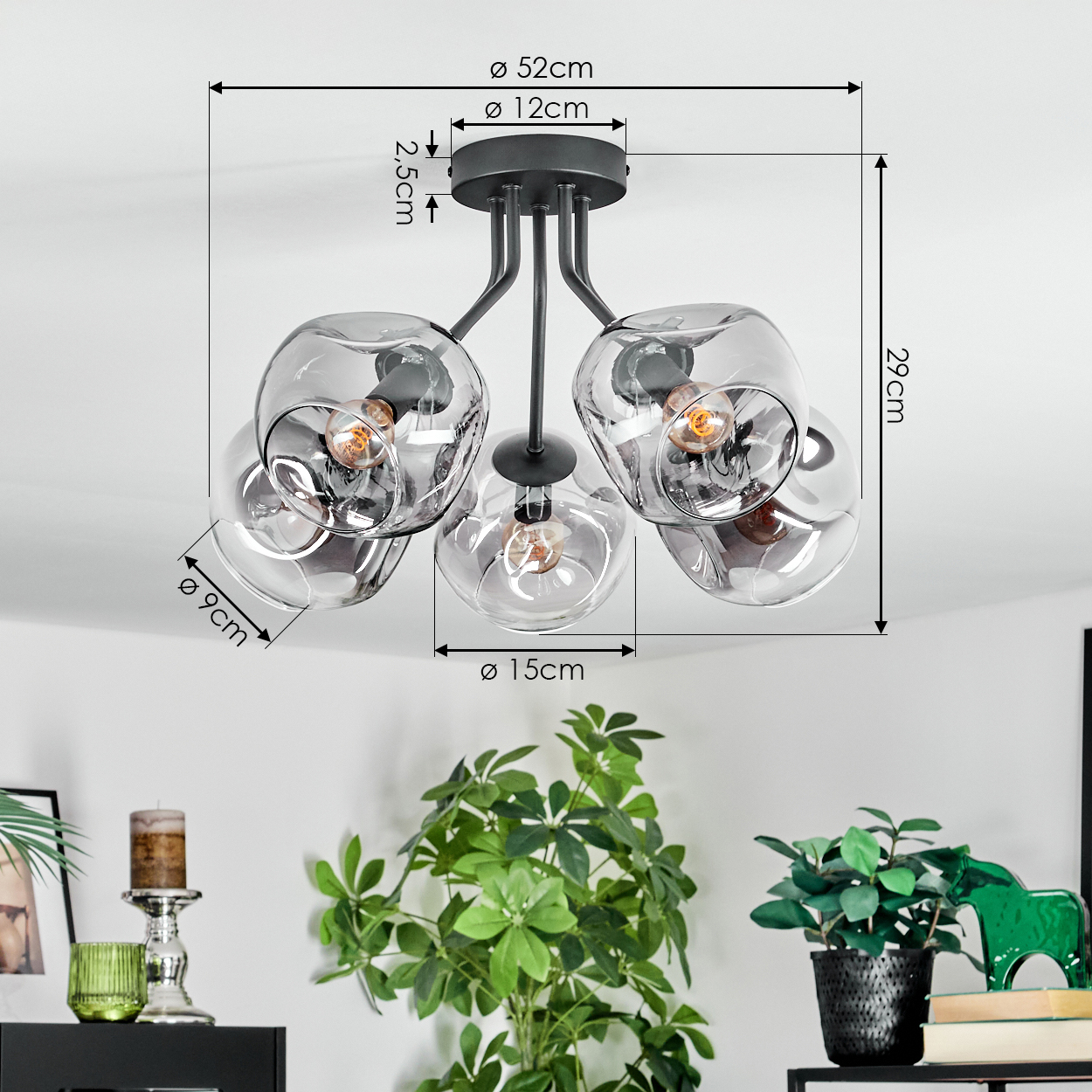 Moderne Deckenlampe mit fünf Glaskugeln, Maße beschriftet. Grüne Pflanzen und ein Kerzenhalter im Vordergrund.