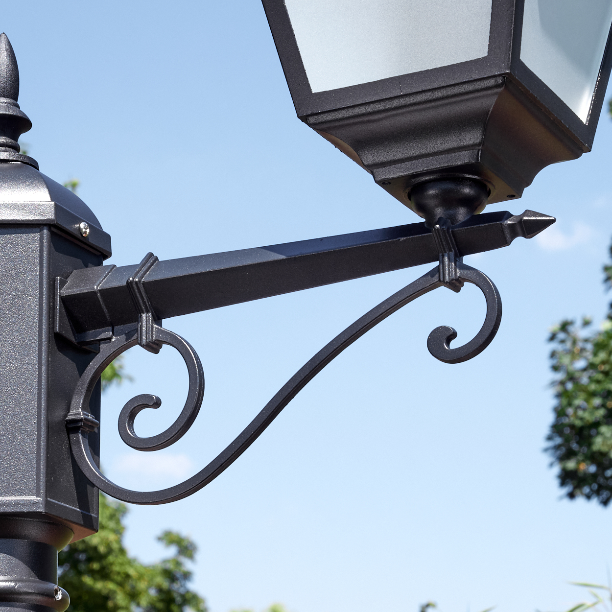 Nahaufnahme einer schwarzen, verzierten Laterne mit dekorativem Arm, Lampe und blauem Himmel im Hintergrund mit grünen Bäumen.