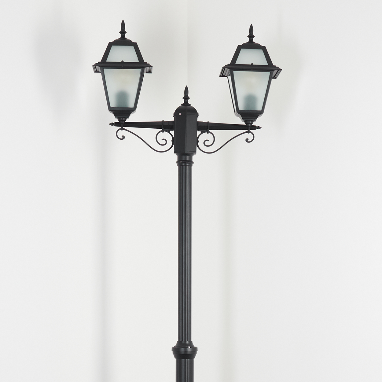 Eine schwarze Straßenlaterne mit zwei Lichtern steht vor einer weißen Wand. Die Lampe hat verzierte Details und Milchglas.