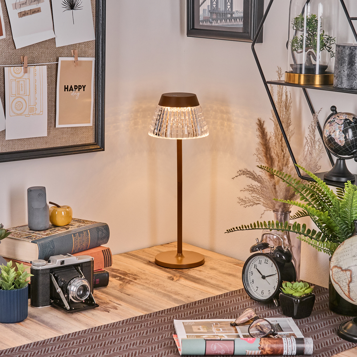 Eine beleuchtete Tischlampe auf einem Holztisch mit Büchern, einer Kamera und einer Uhr. Dekoartikel im Hintergrund auf einem Regal.