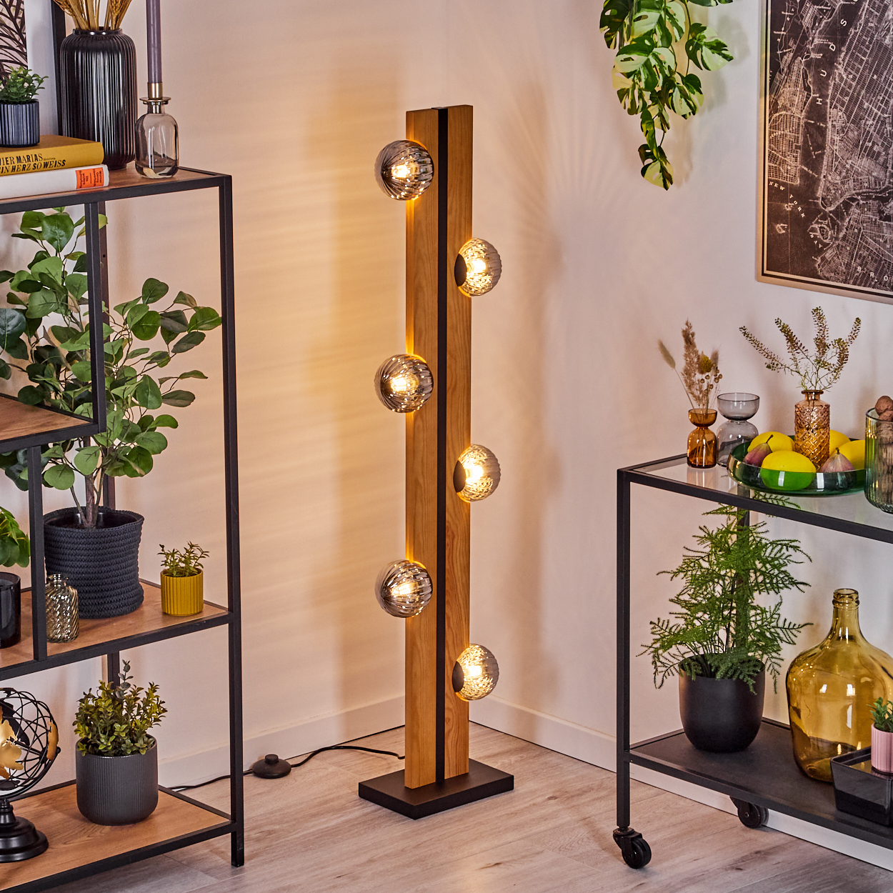 Eine moderne Stehlampe mit fünf Glaskugeln steht in einem Raum mit Regalen, Pflanzen und einer schwarz gerahmten Karte. Die Lampe ist aus Holz.