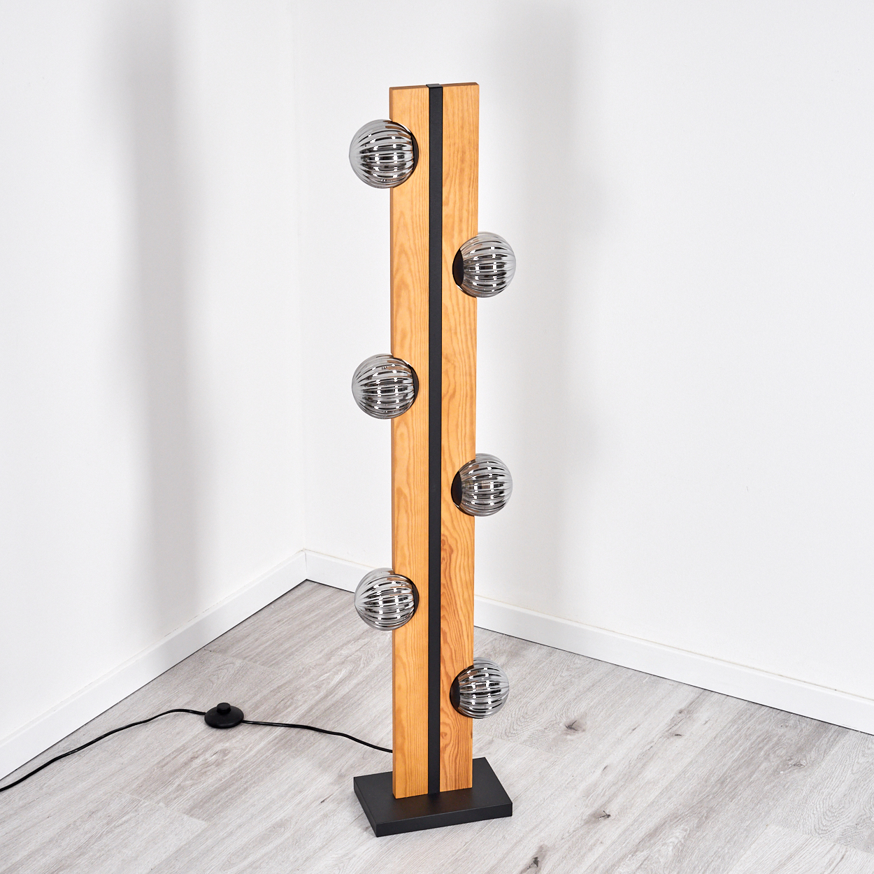 Holz-Stehlampe mit sechs kugelförmigen Lichtern. Sie hat einen schwarzen Sockel und Streifen und steht in einer Ecke mit weißen Wänden.