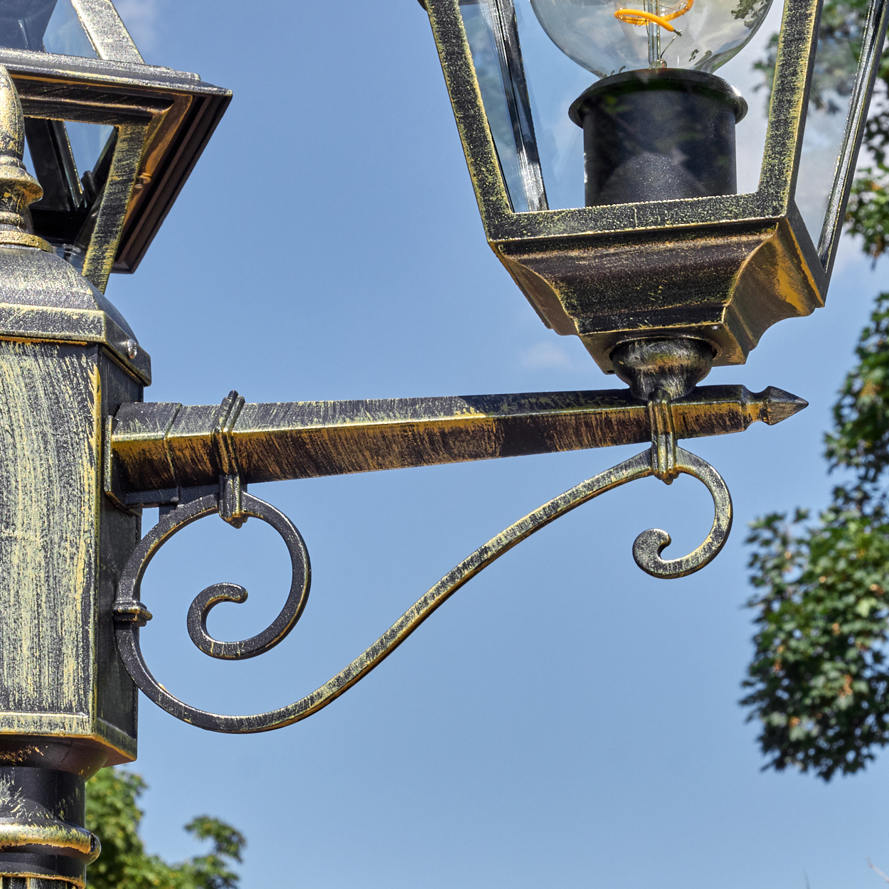 Nahaufnahme einer dekorativen schwarz-goldenen Straßenlaterne vor blauem Himmel. Eine Glühbirne ist im Glaskasten der Lampe zu sehen.