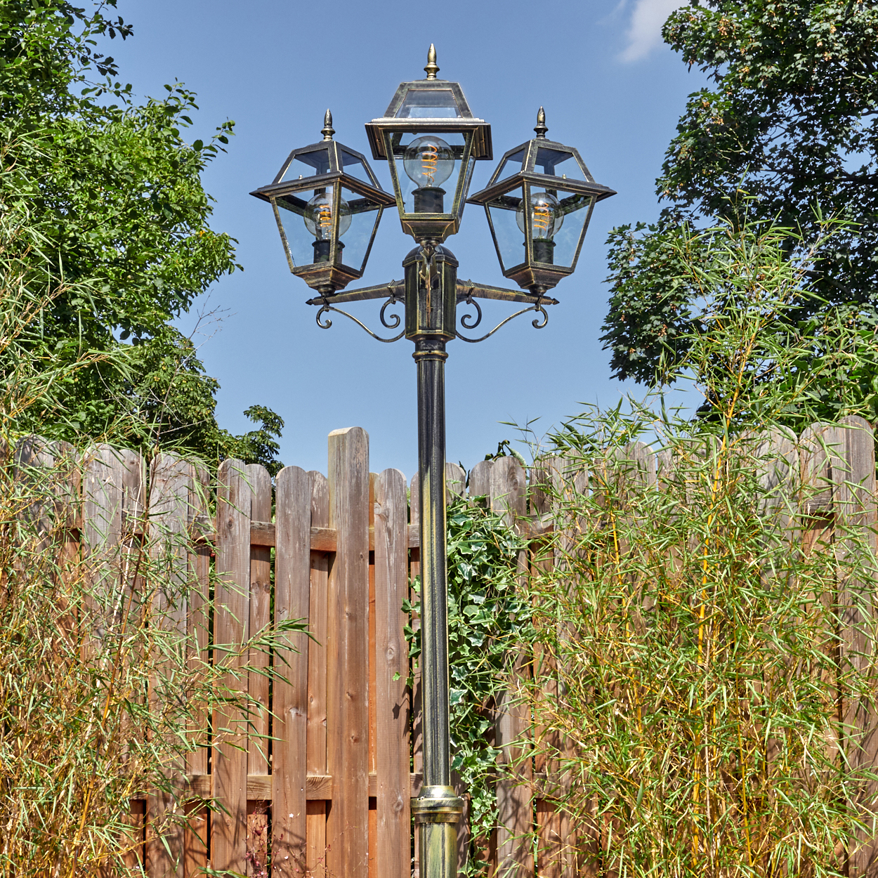 Eine dekorative Laterne mit drei Lampen steht vor einem Holzzaun. Grüne Pflanzen und blauer Himmel sind im Hintergrund.