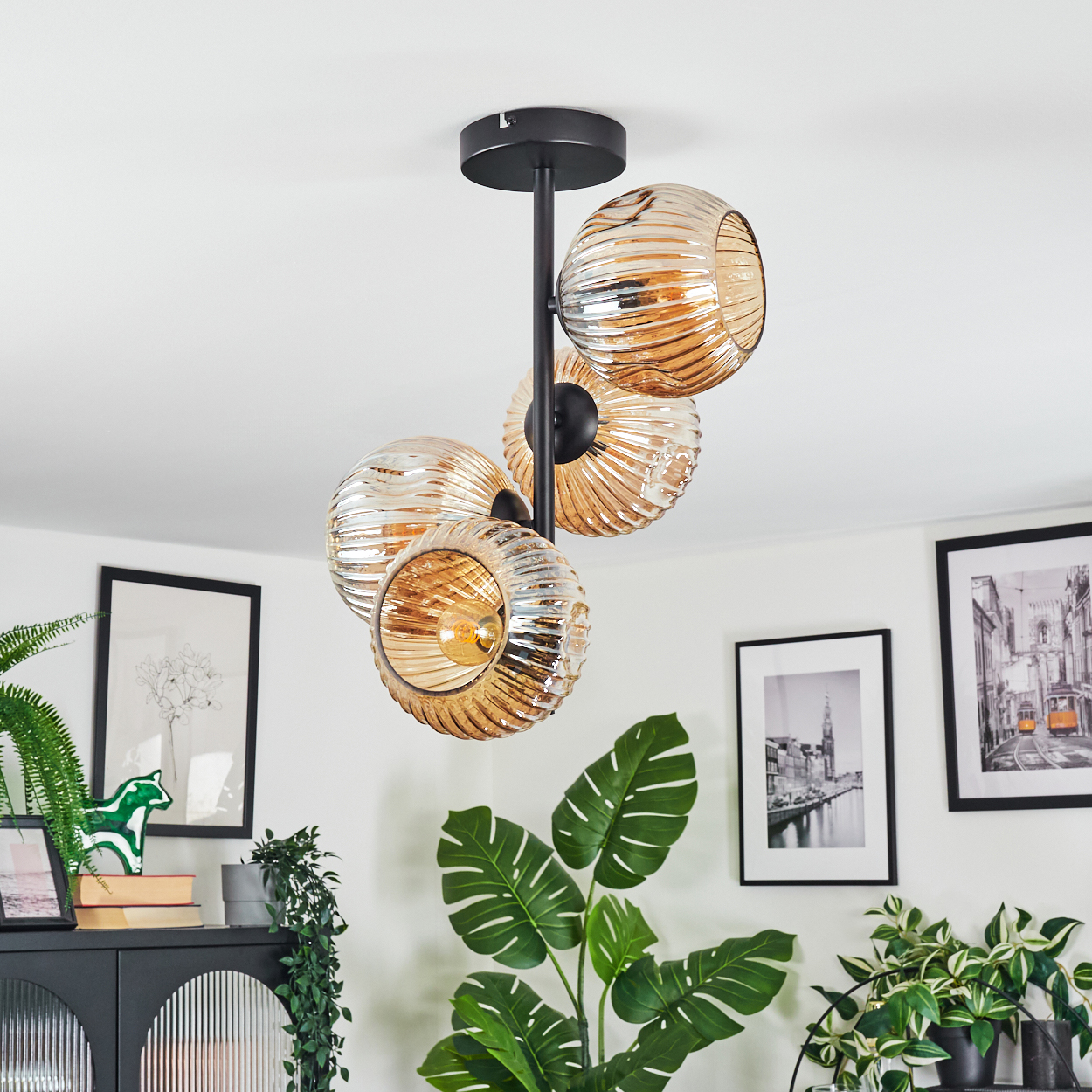 Eine moderne Deckenlampe mit vier Glaskugeln hängt. Darunter dekorieren Pflanzen und gerahmte Kunst den Raum. Die Decke ist weiß.