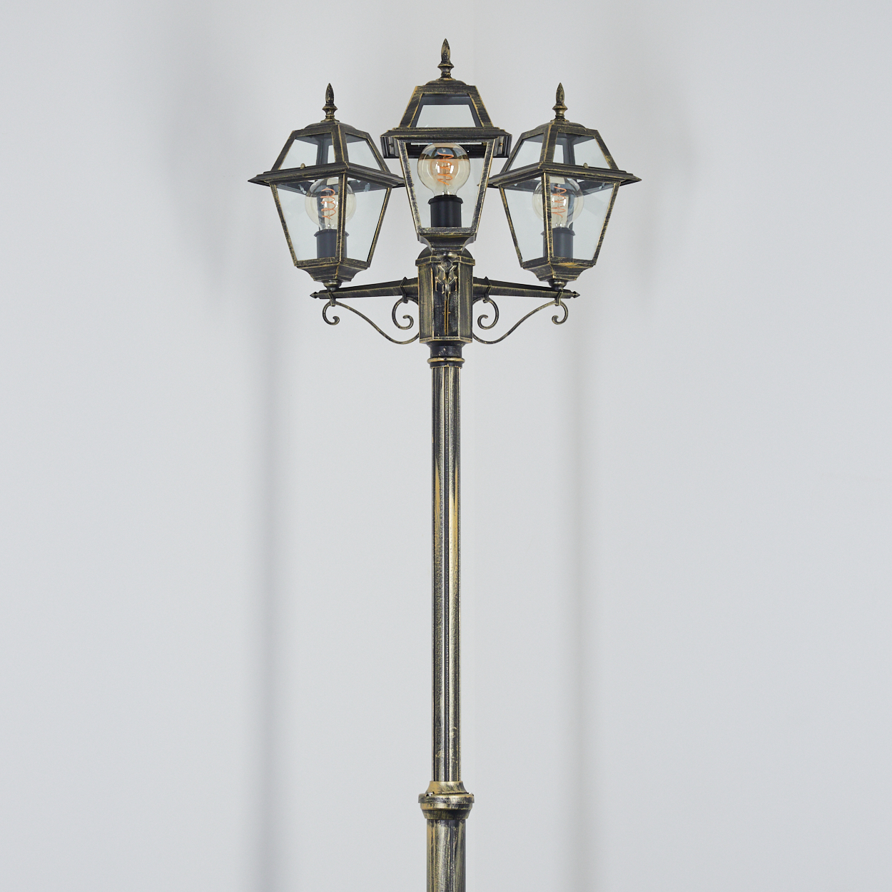 Eine verzierte Laterne mit drei Lampen und Glasgehäusen. Der Pfosten ist golden und steht vor einer weißen Wand.