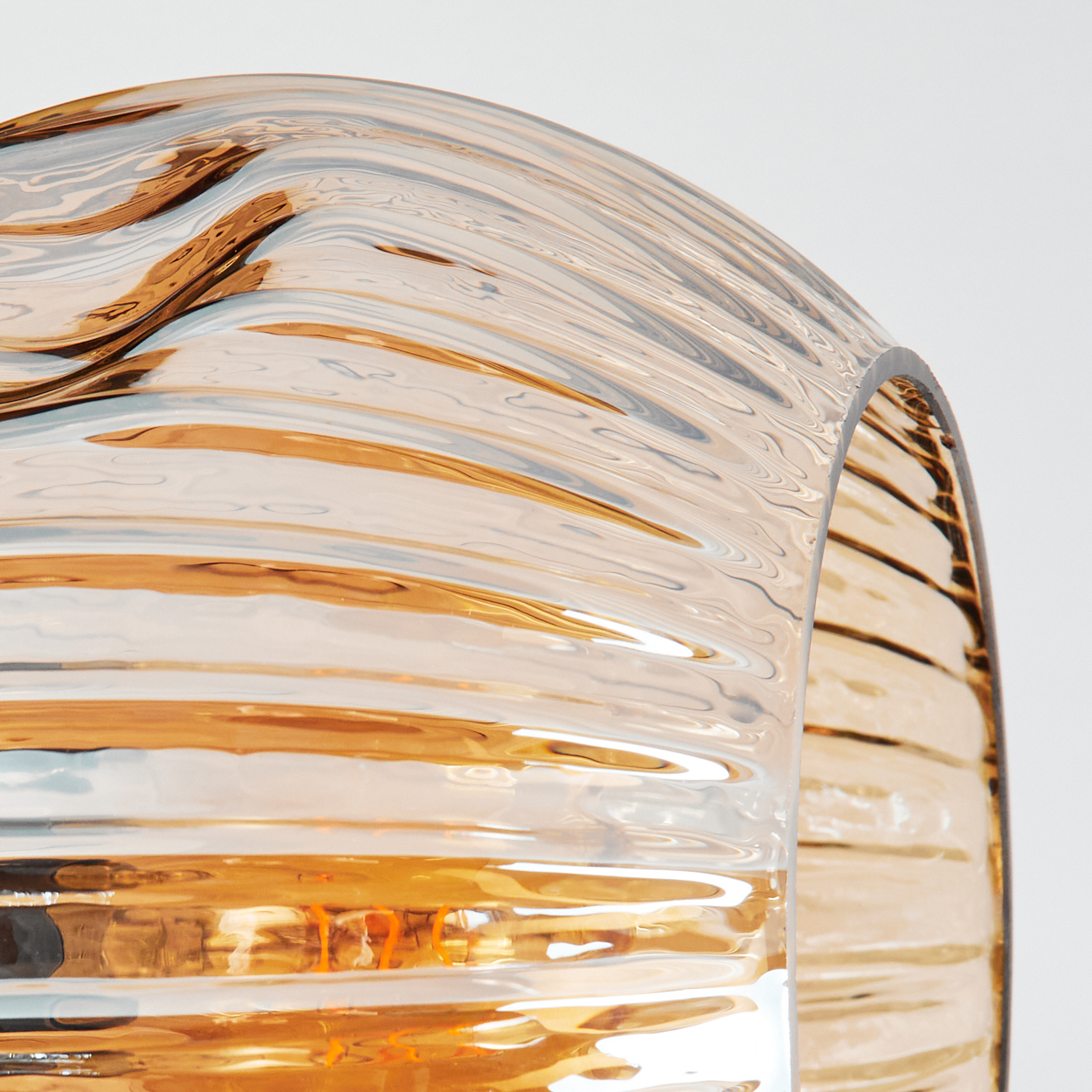 Nahaufnahme eines gebogenen Glasobjekts. Das Glas ist bernsteinfarben mit horizontalen Rippen, die Licht vor weißem Hintergrund reflektieren.