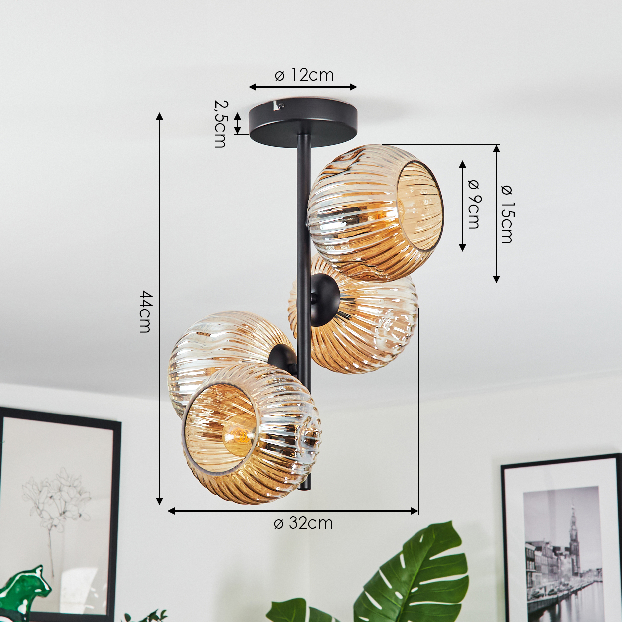 Eine moderne Deckenlampe mit vier bernsteinfarbenen Glaskugeln. Die Lampe ist schwarz und hat in cm beschriftete Abmessungen. Der Hintergrund enthält Kunst und Pflanzen.