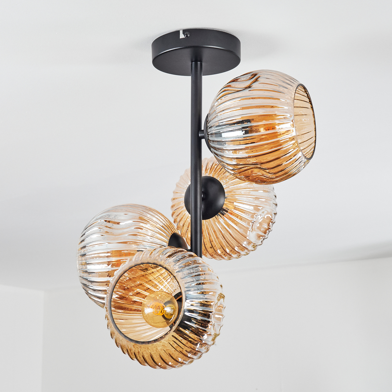 Eine moderne Deckenlampe mit vier bernsteinfarbenen Glaskugeln. Die Lampe hat einen schwarzen Metallrahmen und befindet sich an einer weißen Decke.
